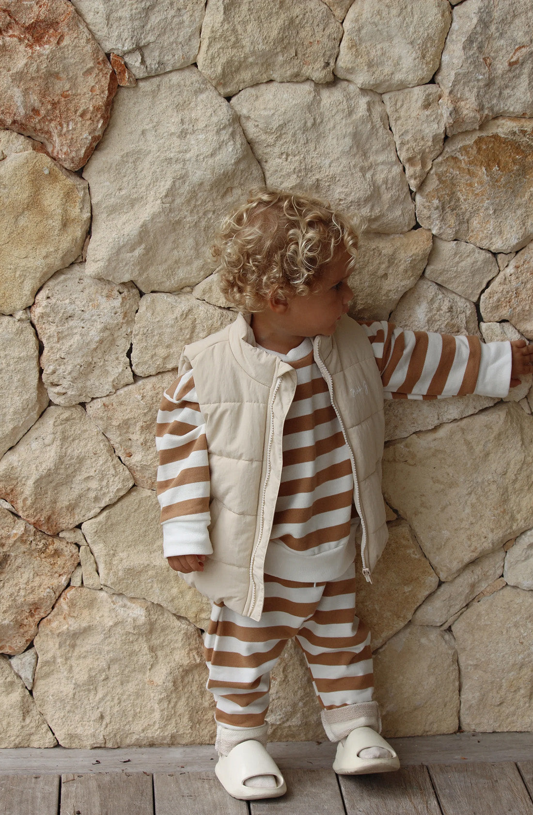 A toddler wearing the Ari Puffer Vest in eggshell over the top of a matching brown and white striped jumper and track pant set. He is standing against a pebbled stone wall with his head facing away from the camera.