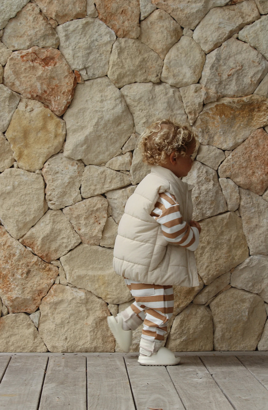 A toddler wearing the ari puffer vest in eggshell over a matching striped brown and white jumper and pant set against a pebbled stone background. He is walking along a wooden deck.