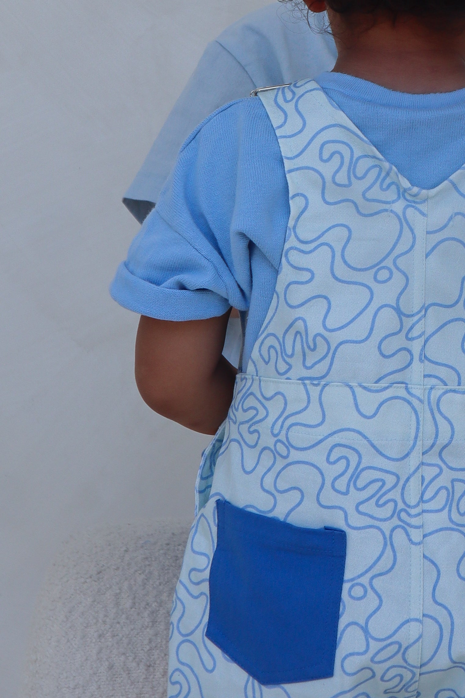 A young child is wearing ZIGGY LOU's Short Overalls Zen, featuring wavy lines and a blue pocket while standing partially facing away. The child embodies a sense of zen design against the plain background.