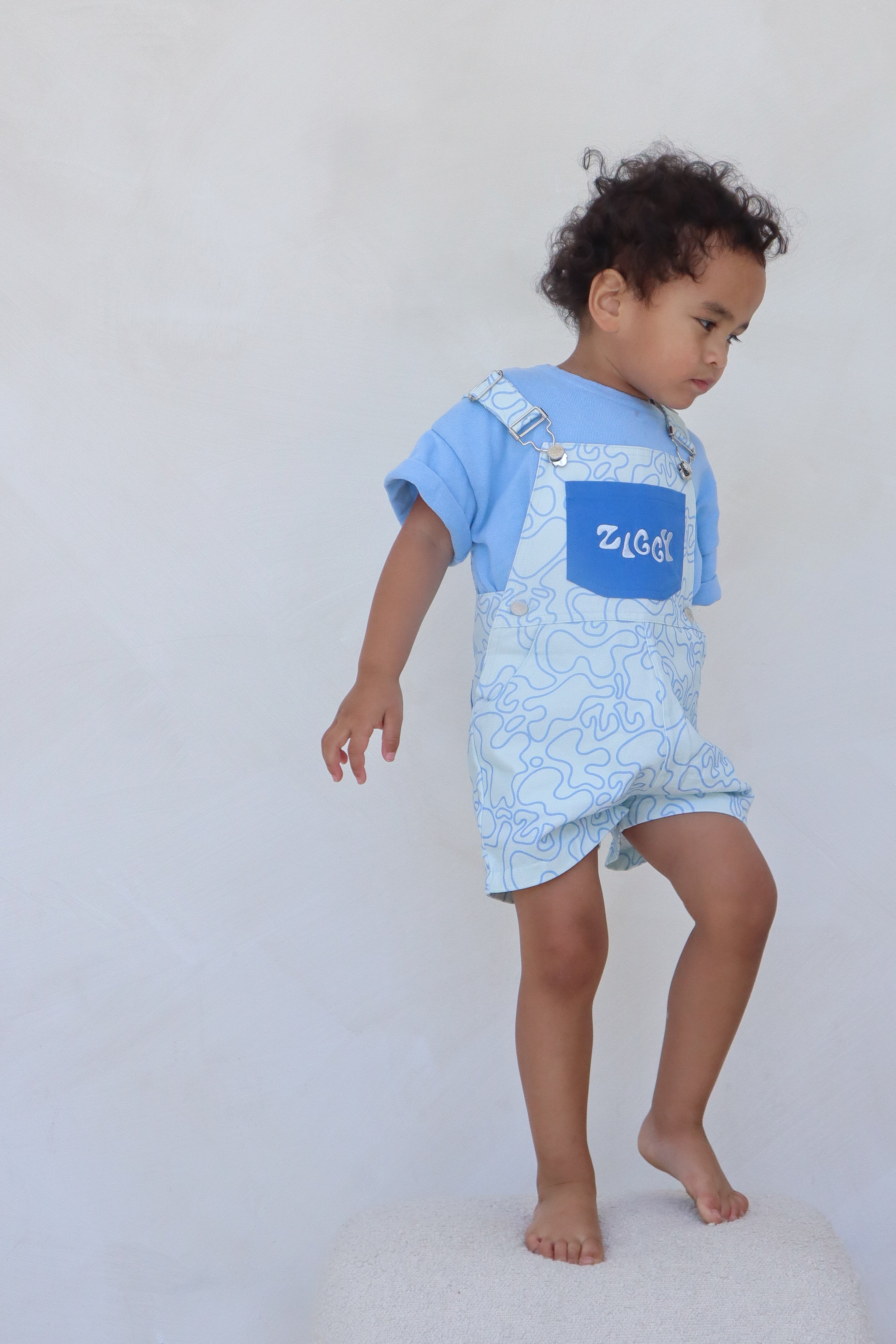 A young child with curly hair stands on a stool against a minimalist, zen-inspired background. They are wearing a light blue t-shirt paired with the ZIGGY LOU Short Overalls Zen, made from 100% cotton canvas in a patterned blue and white design. The child is looking down with one foot slightly raised.