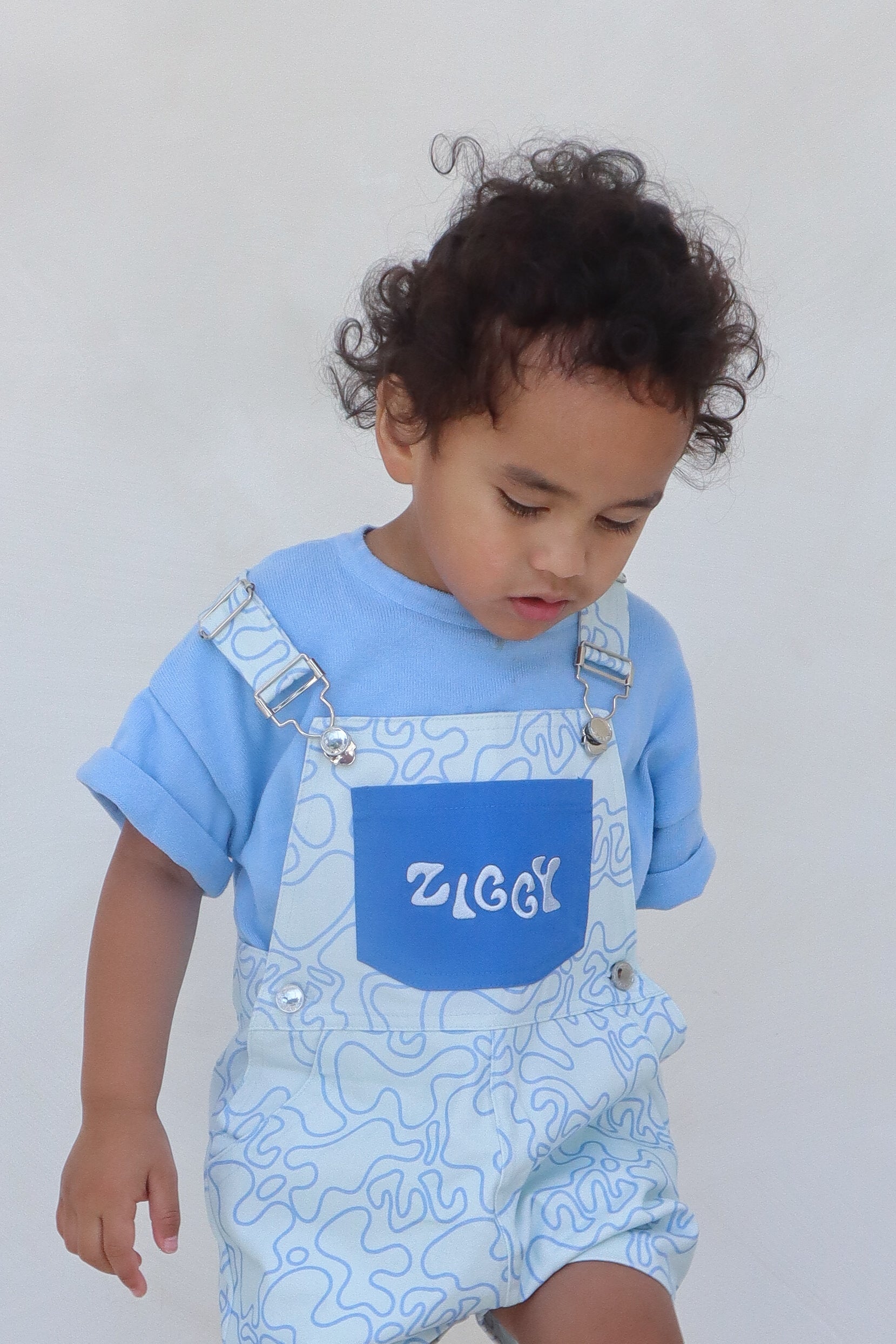 A toddler with curly hair is wearing a light blue shirt under the Short Overalls Zen, made from 100% cotton canvas by ZIGGY LOU, with "ZIGGY" on the front. The child gazes down against a plain white background.