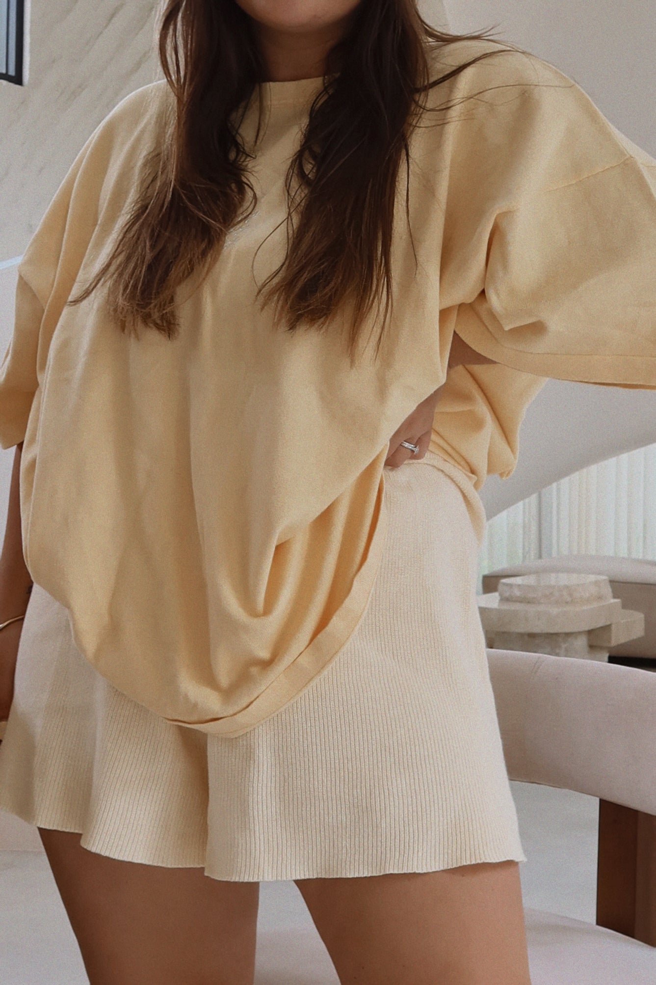 A person wearing an oversized ZIGGY LOU Women's Signature Tee in Butter and matching shorts stands in a modern, minimalist room with neutral-colored furniture. Their hand rests on their hip, and long hair cascades over their shoulders.