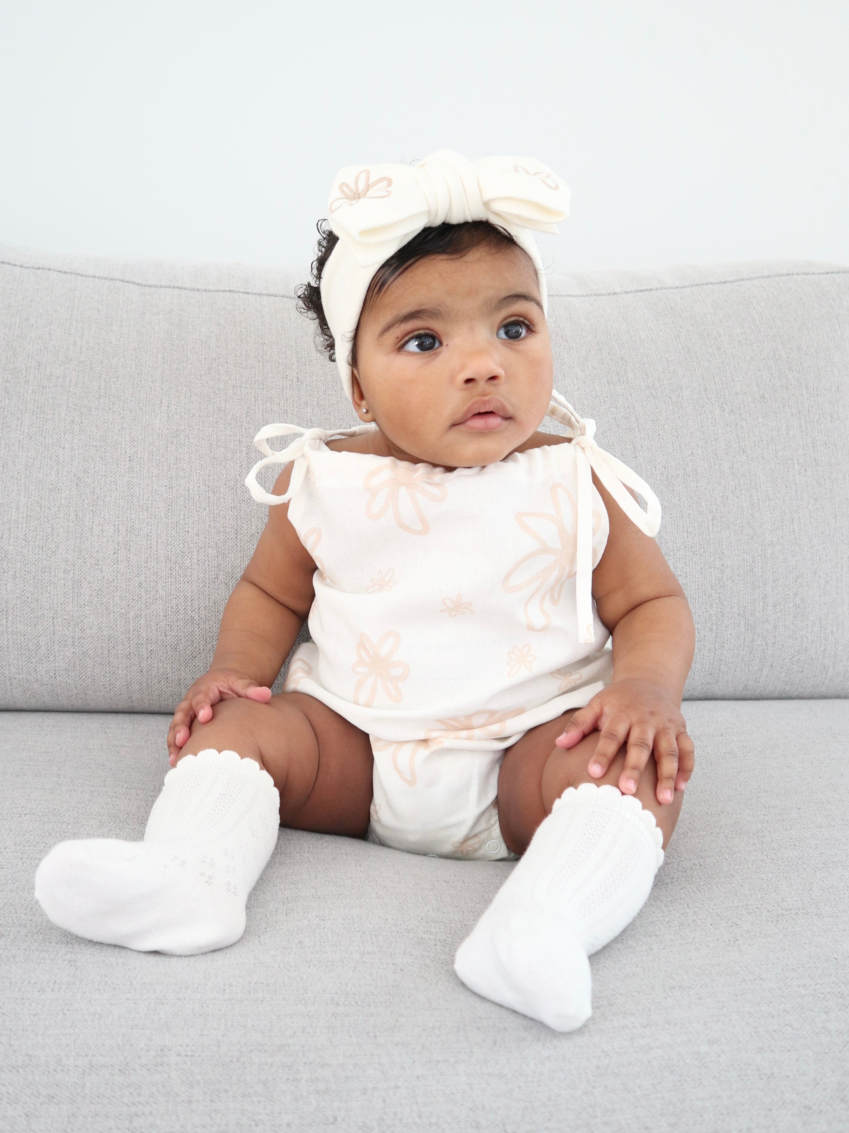 A baby dressed in the Eva Bubble Romper Gia by ZIGGY LOU, adorned with floral patterns, is seated on a gray couch. The ensemble includes a coordinating headband with a large bow and white socks.