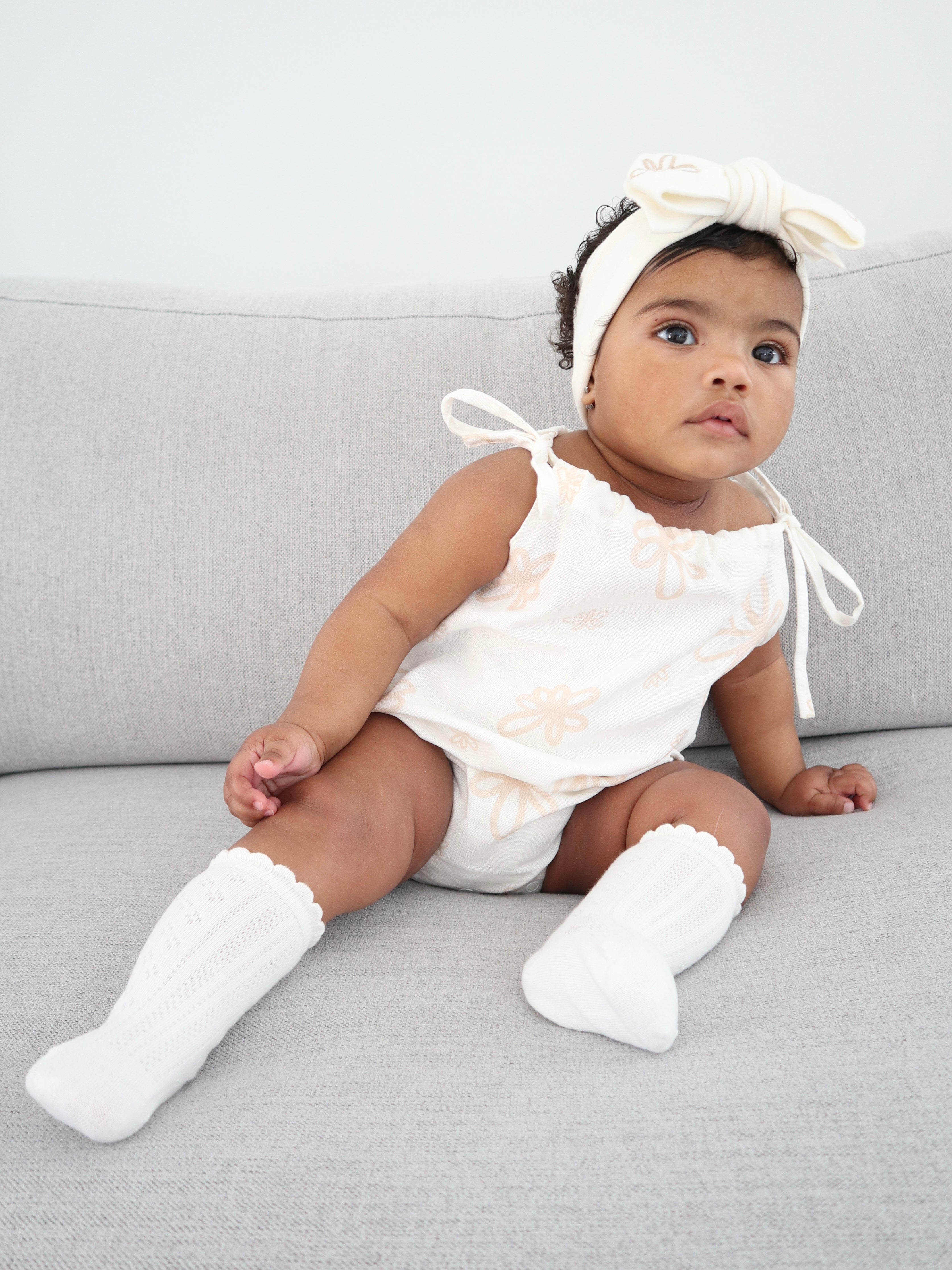 A baby sits on a gray sofa, dressed in an Eva Bubble Romper Gia from ZIGGY LOU, paired with matching knee-high socks and a headband adorned with a bow. The lightweight outfit complements the baby's curious expression as they gaze upwards.