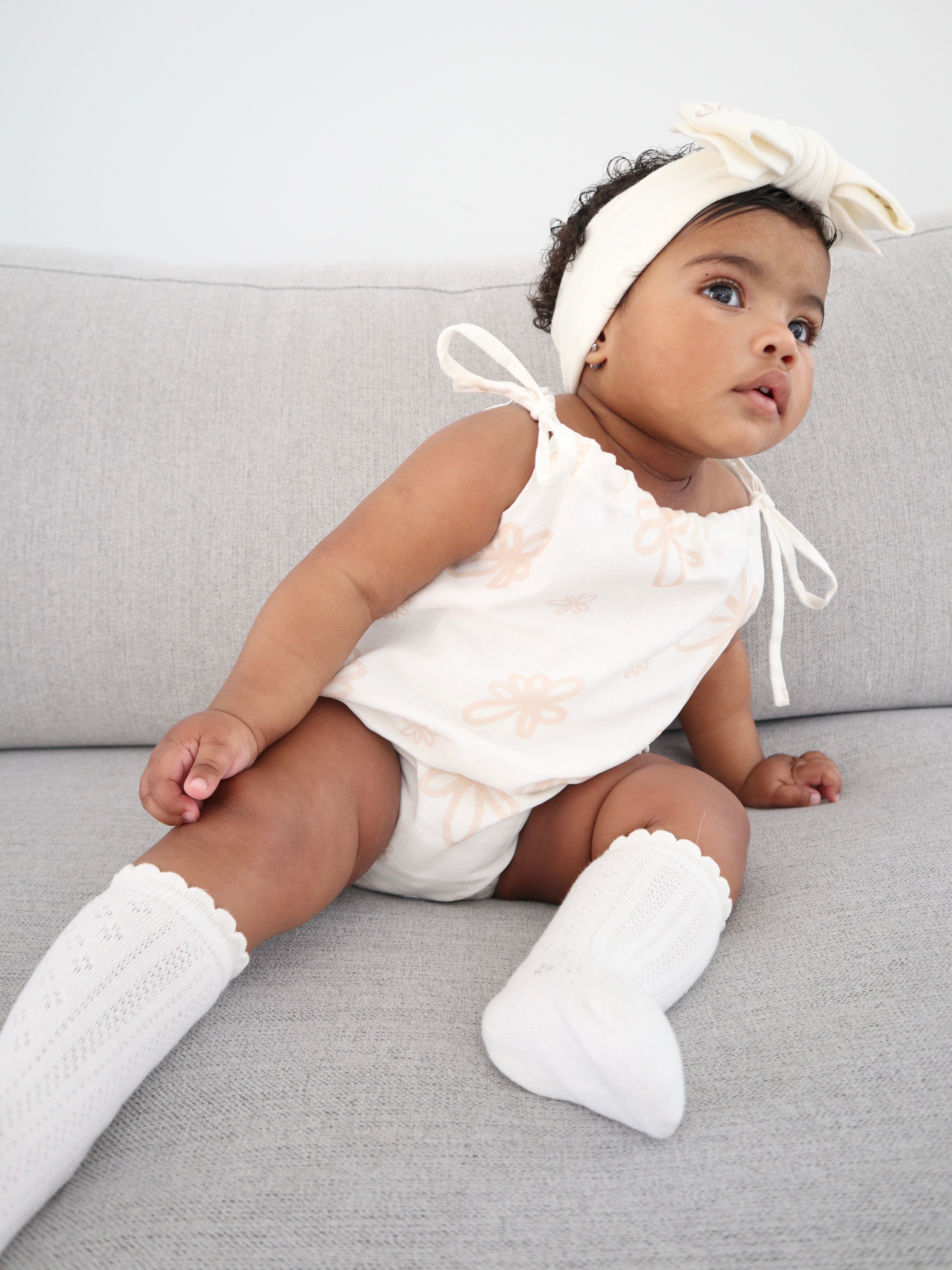 A baby, wearing ZIGGY LOU's Lace Socks Milk in a white floral outfit with custom-dyed lace accents and matching knee-high socks, sits on a light gray sofa. The headband is accented by a large bow as the baby gazes curiously to the side.