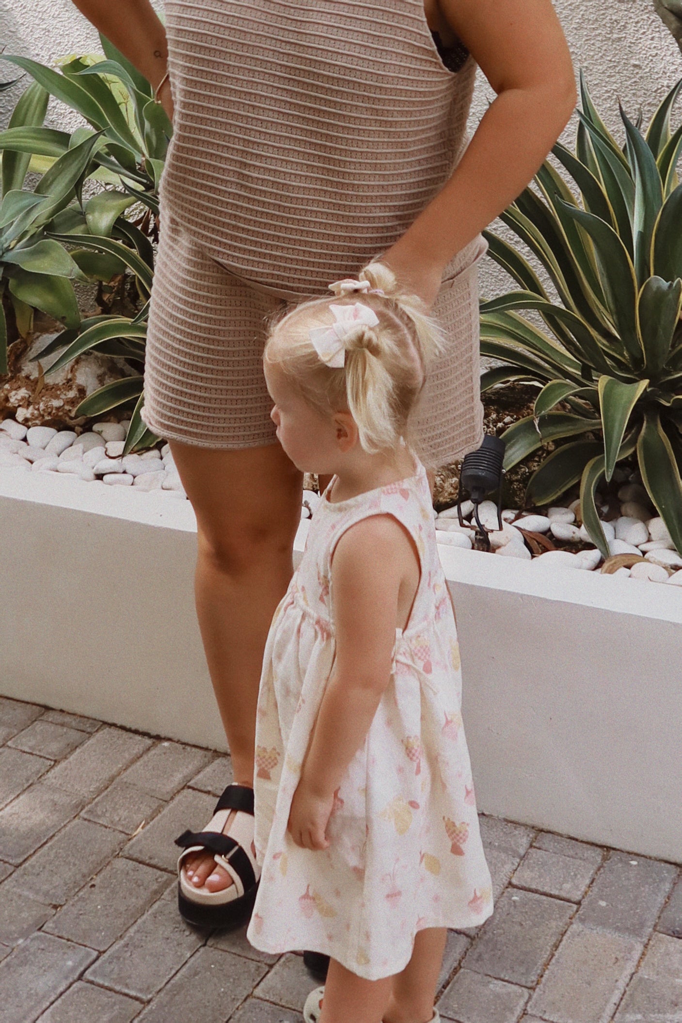 A toddler wearing the Sofia Dress August from ZIGGY LOU, adorned with a bow in her hair, stands next to a woman dressed in a textured brown ensemble. They are on a paved path with decorative plants and white stones in the background.