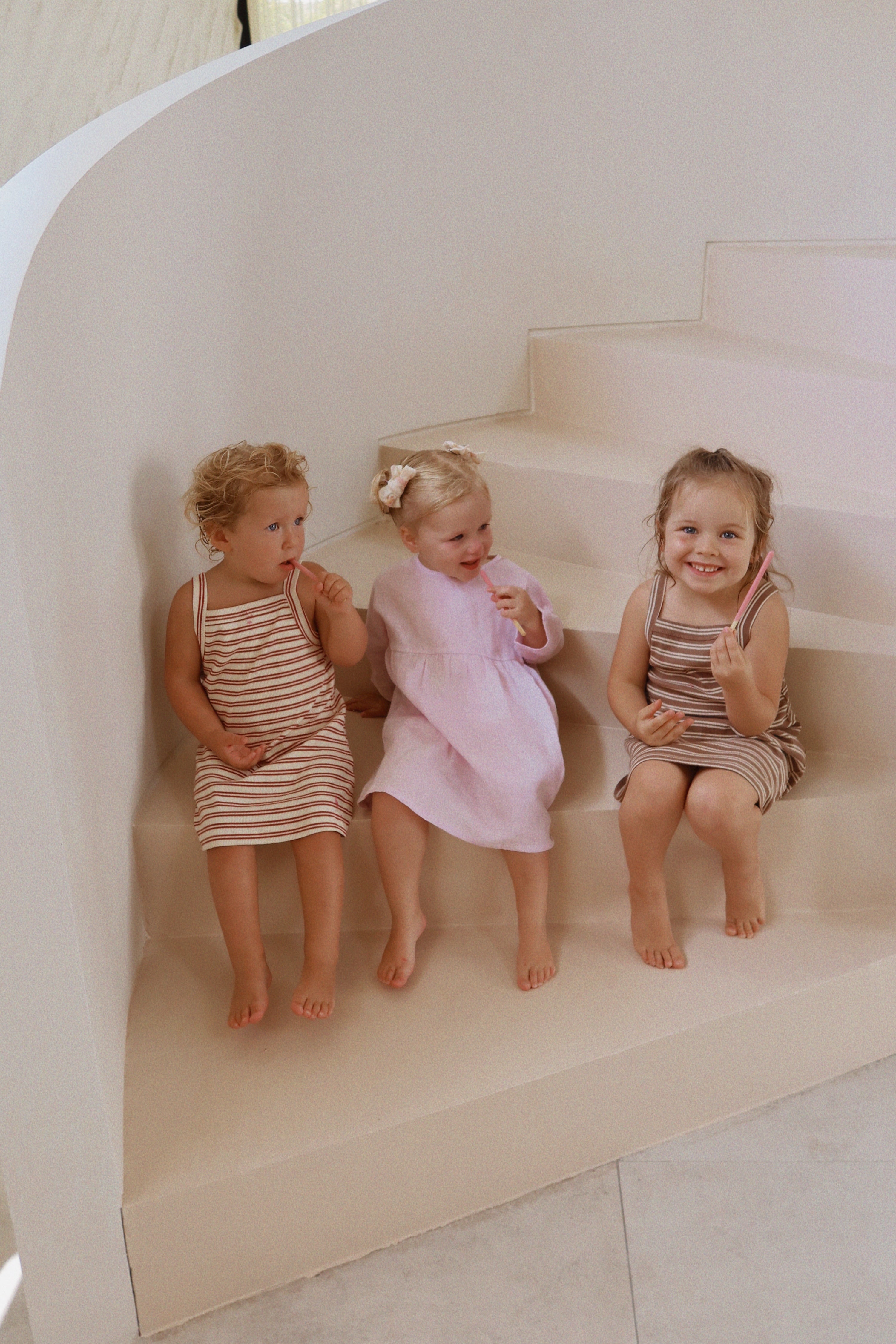 Three young children sit on light-colored stairs. Two wear matching knee-length stripped dresses by MILKY DESIGNS, while the third wears a pink Posy Dress Lilac, Size 5. All are barefoot and smiling in a bright, minimalistic setting holding small items.