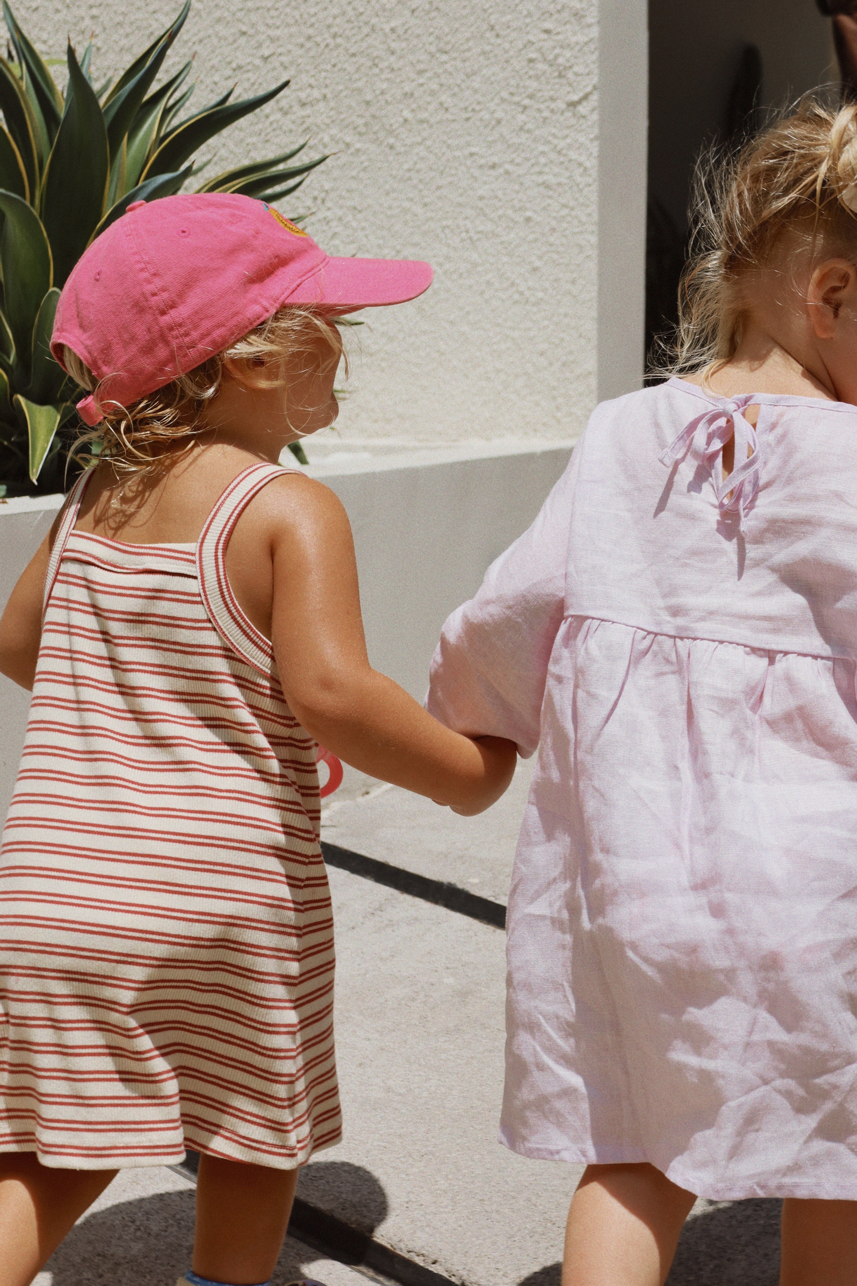 Two young children stroll together, hand in hand. One is dressed in the Posy Dress Lilac by MILKY DESIGNS, featuring adjustable ties, while the other wears a chic striped dress paired with a pink cap. They enjoy a sunny day outdoors, enveloped by lush greenery and set against the backdrop of a white building.