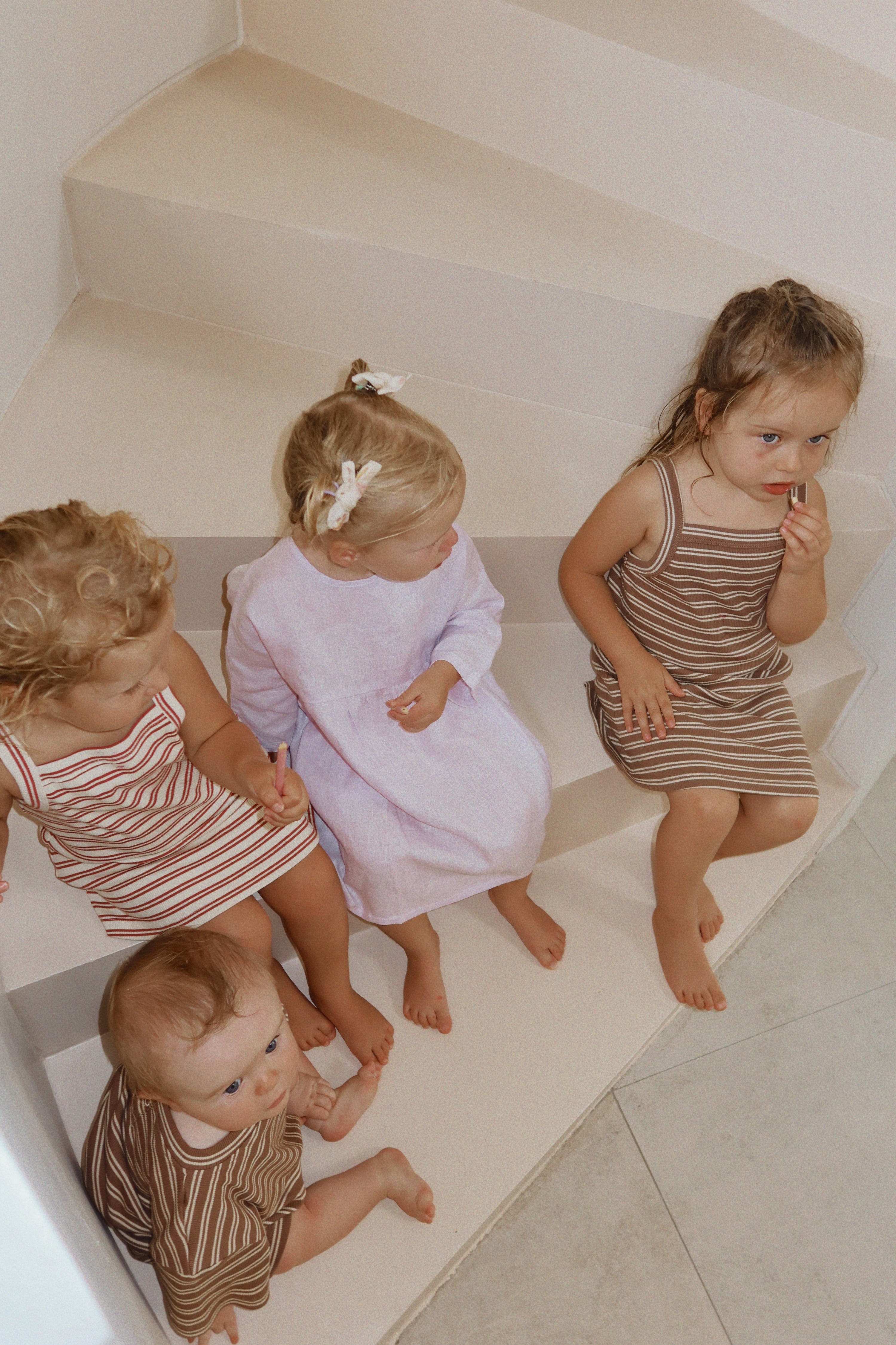 Four young children sit on light-colored stairs. Three wear knee-length striped outfits in brown and white linen, while the fourth wears a Posy Dress Lilac by MILKY DESIGNS. Featuring adjustable ties, they appear engaged and curious, with one child looking upwards.