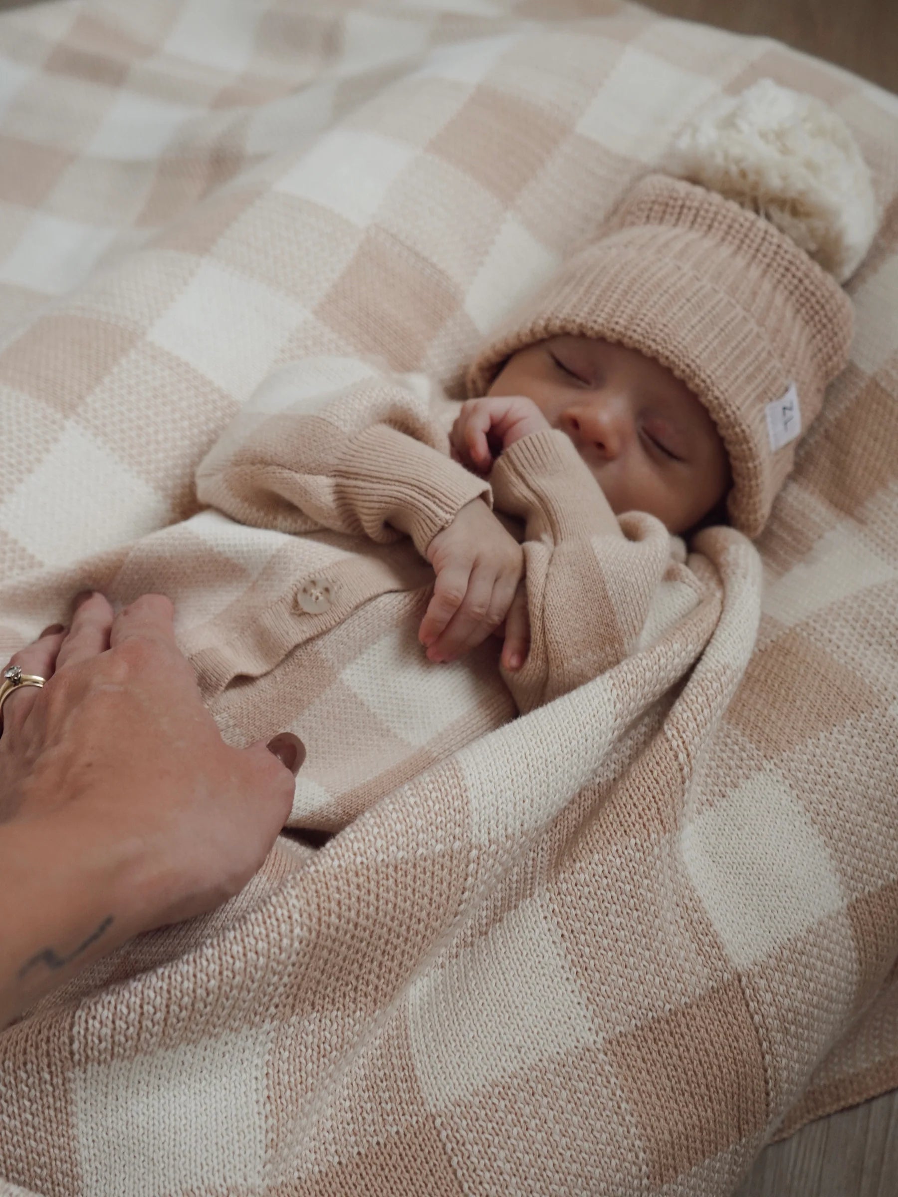A sleeping baby in a beige knit outfit and hat lies on the Ziggy Lou - Blanket Gingham by ZIGGY LOU. An adult hand gently rests on the baby’s chest, reflecting tenderness and a focus on newborn safety.