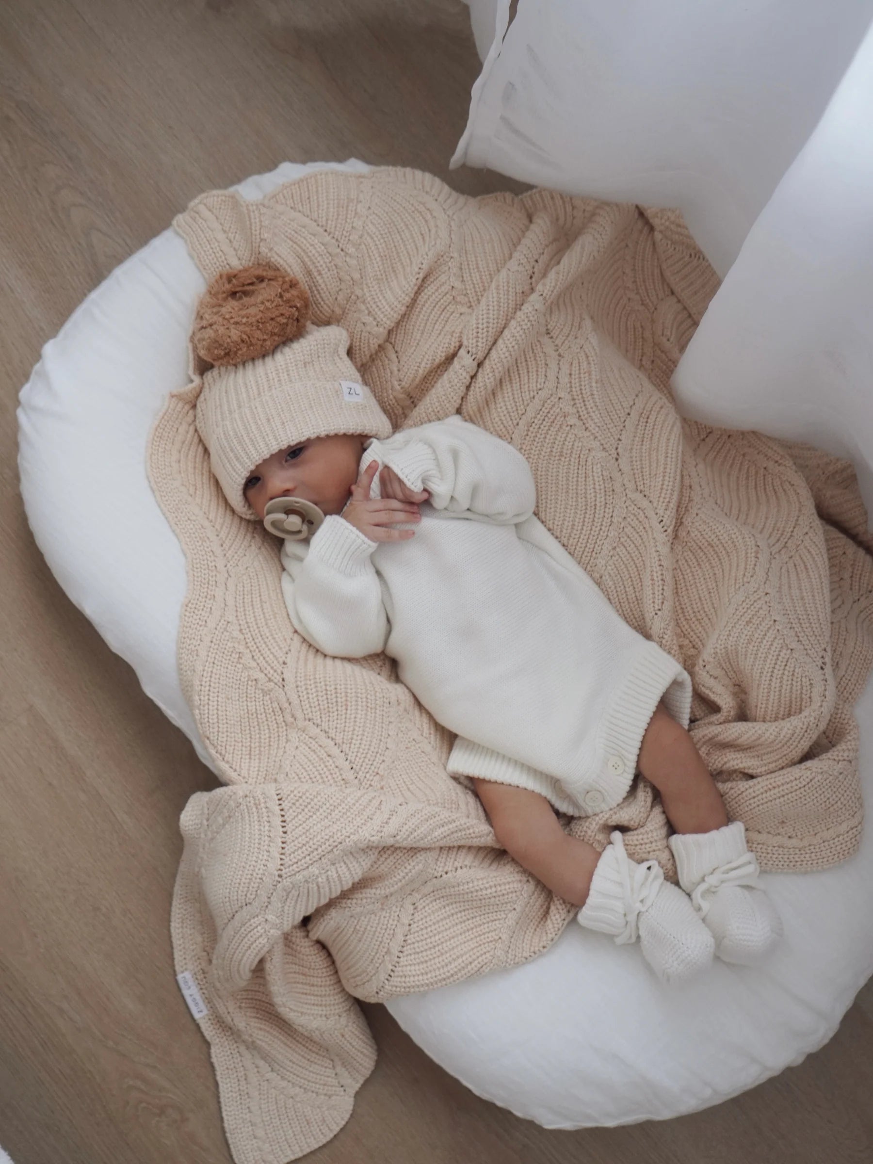 A baby in a cream cotton knit outfit, hat with pom-pom, and booties lies on the ZIGGY LOU Ziggy Lou - Blanket Biscotti Fleck in a white cushioned nest on a wooden floor, holding a pacifier and looking up.