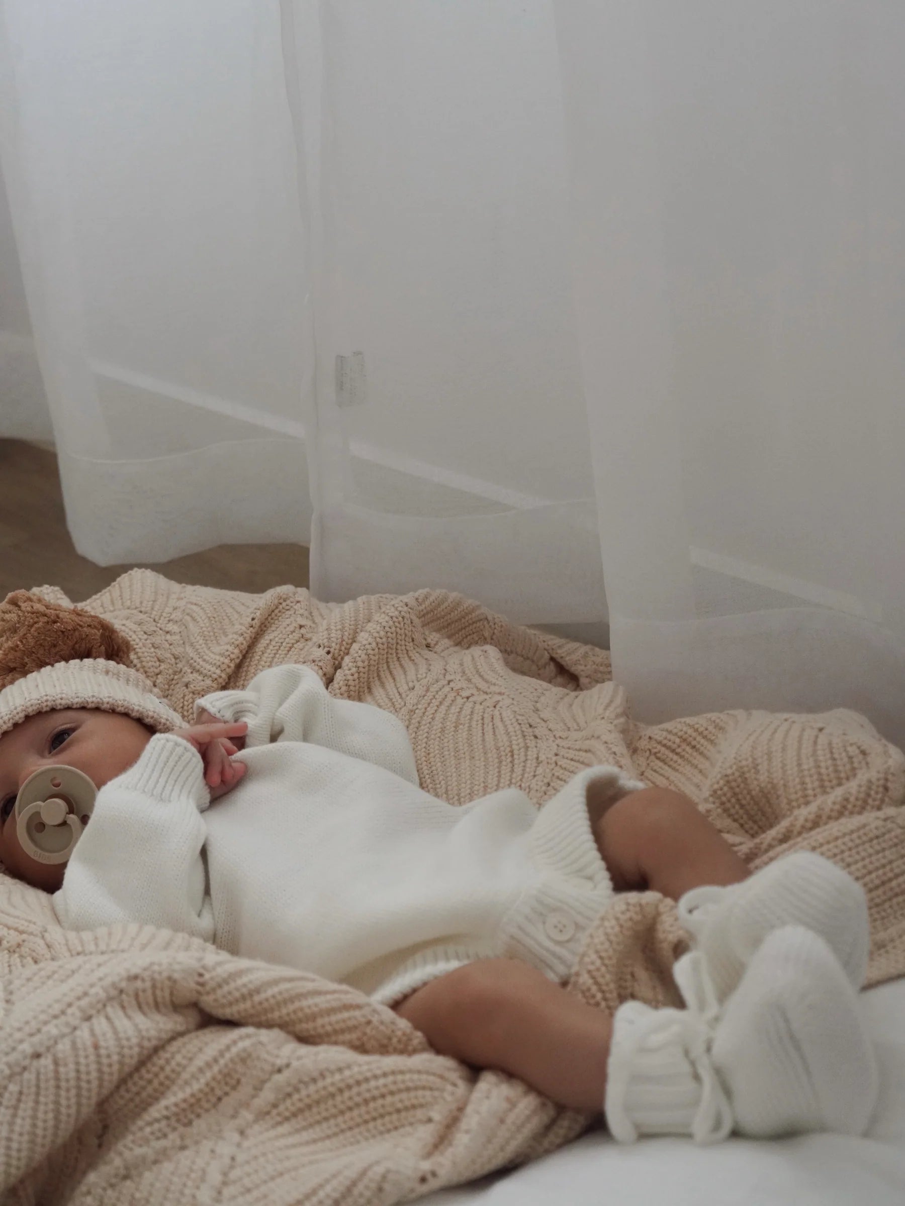 A baby wearing a white knit outfit, Ziggy Lou - Booties Milk by ZIGGY LOU, and a hat lies on a beige textured blanket with a pacifier. Soft natural light streams through sheer white curtains in the background.