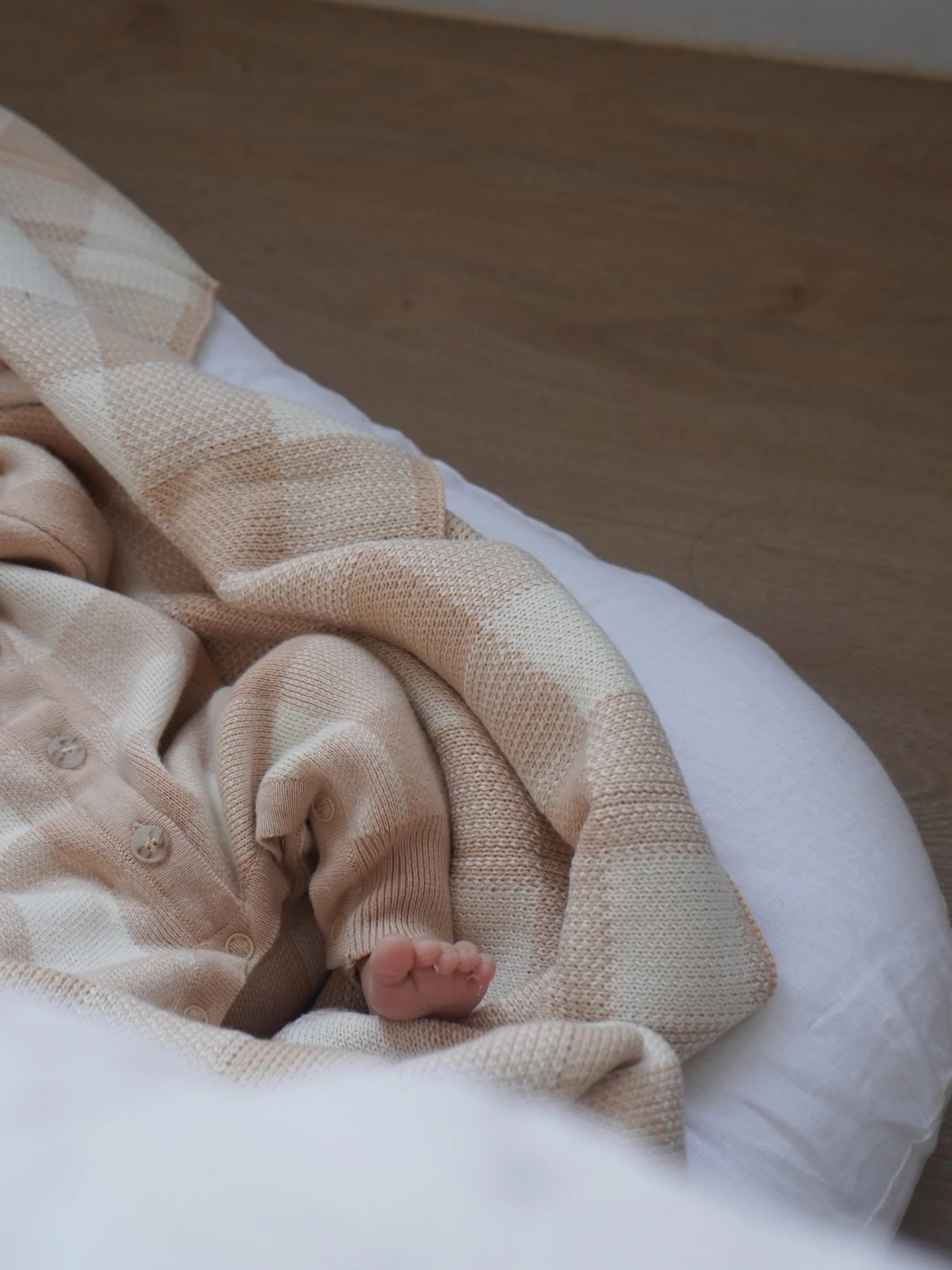 A baby rests on a white bed, wrapped in a soft, machine-washable ZIGGY LOU Blanket Gingham. Only part of the baby's body is visible: a small foot and part of the arm and torso in neutral clothing.