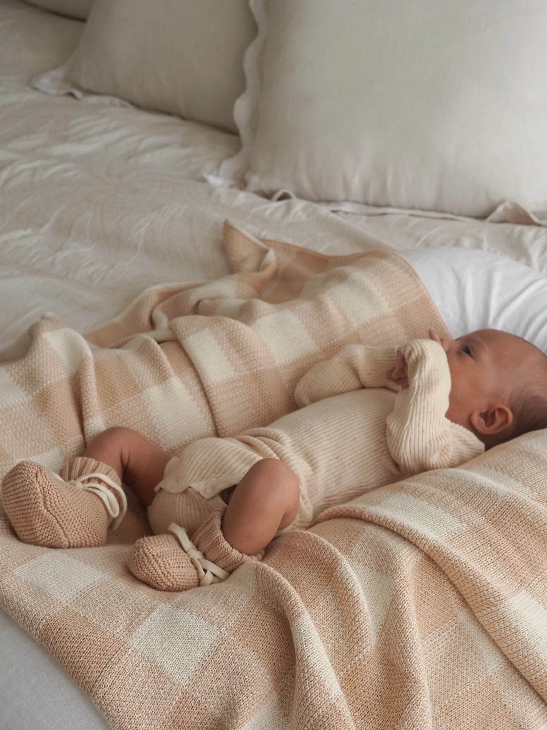 A baby in a beige knitted outfit and booties lies on a ZIGGY LOU Ziggy Lou - Blanket Gingham, a soft, machine washable cotton blanket in beige and white checks, ensuring comfort and safety.