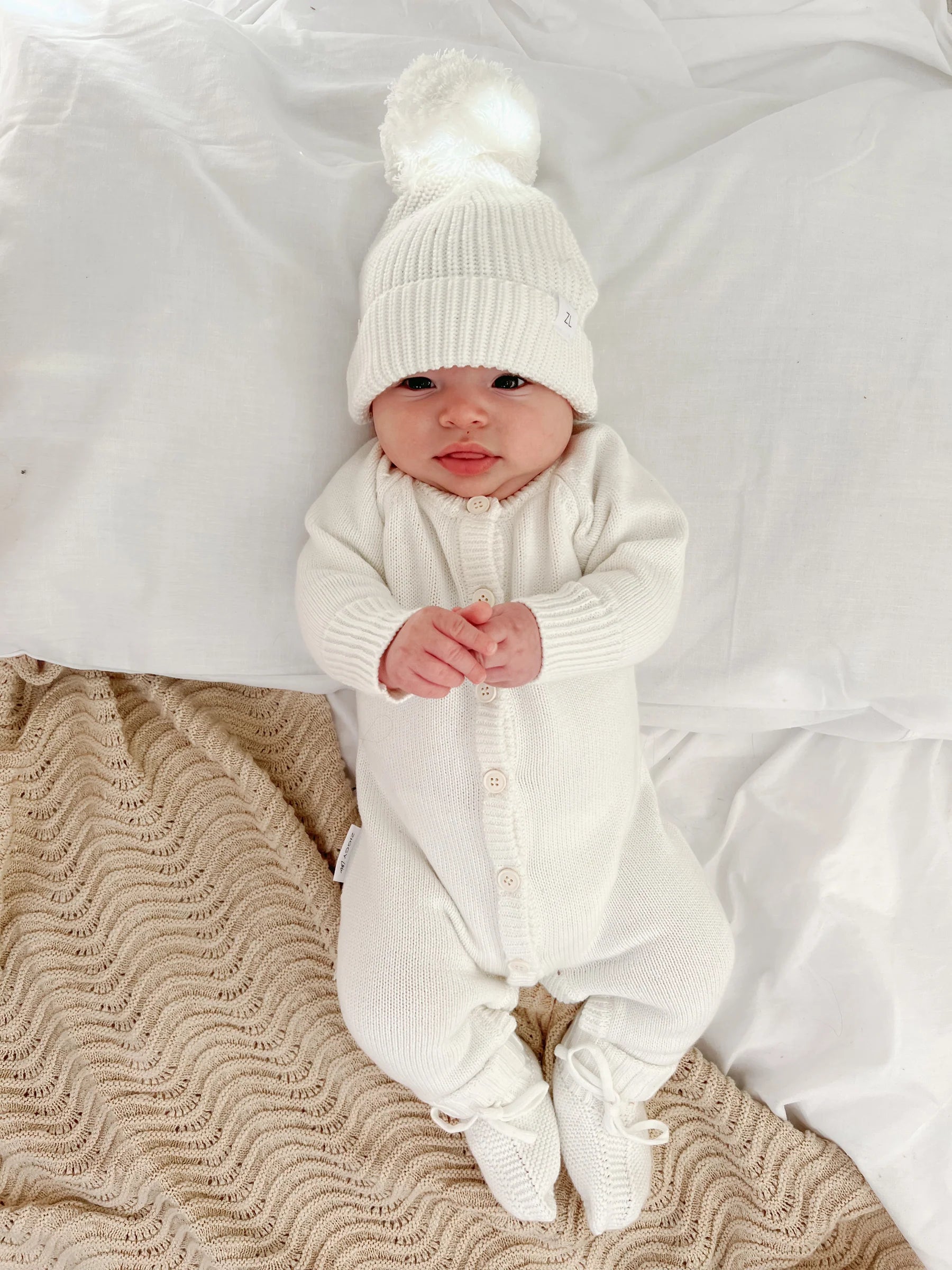 A baby in a ZIGGY LOU Classic Knit Romper Milk lies on a white bed, hands clasped, wearing a matching beanie with a pom-pom and snuggled near a beige textured blanket—a cozy winter look.