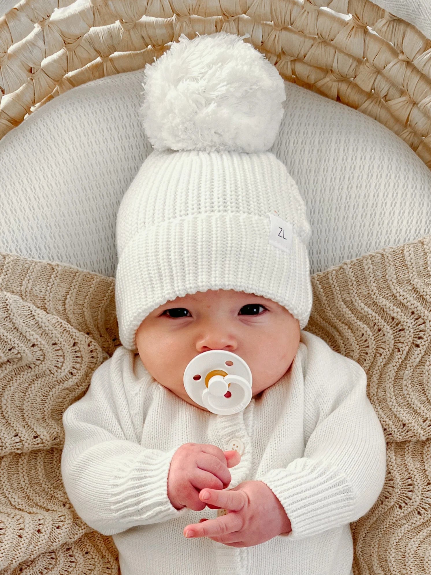 A baby from the Milk Signatures Collection wears a ZIGGY LOU Ziggy Lou - Beanie Milk, a chunky knit beanie with a large pom-pom, paired with a 100% cotton outfit, lying on a textured cream blanket and holding a pacifier.