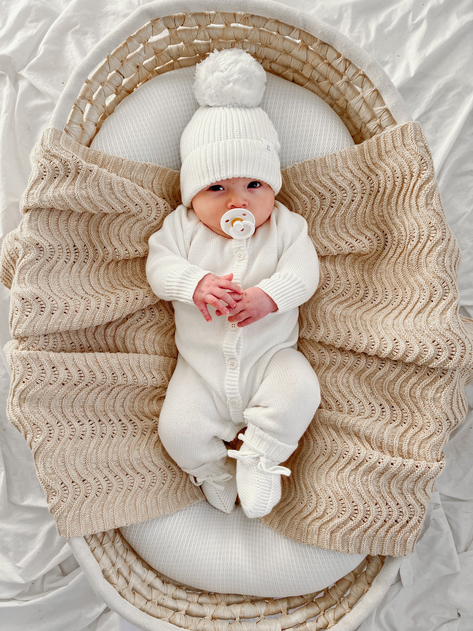 A baby in the Ziggy Lou - Classic Knit Romper Milk by ZIGGY LOU and a pom-pom hat lies in a woven bassinet on a beige textured blanket, holding a pacifier—a cozy scene perfect for showcasing adorable winter baby clothing.