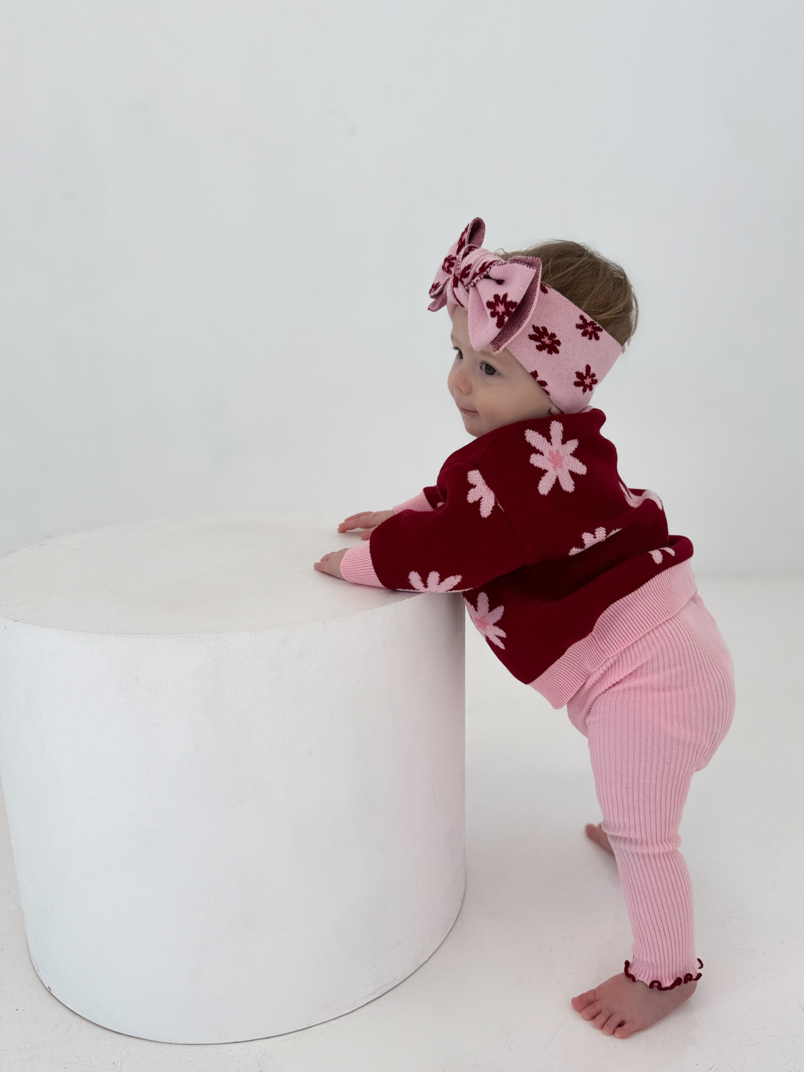 A baby wearing ZIGGY LOU Leggings Lola, a red sweater with pink flowers, and a matching headband stands barefoot, holding a white cylindrical object against a plain white background.