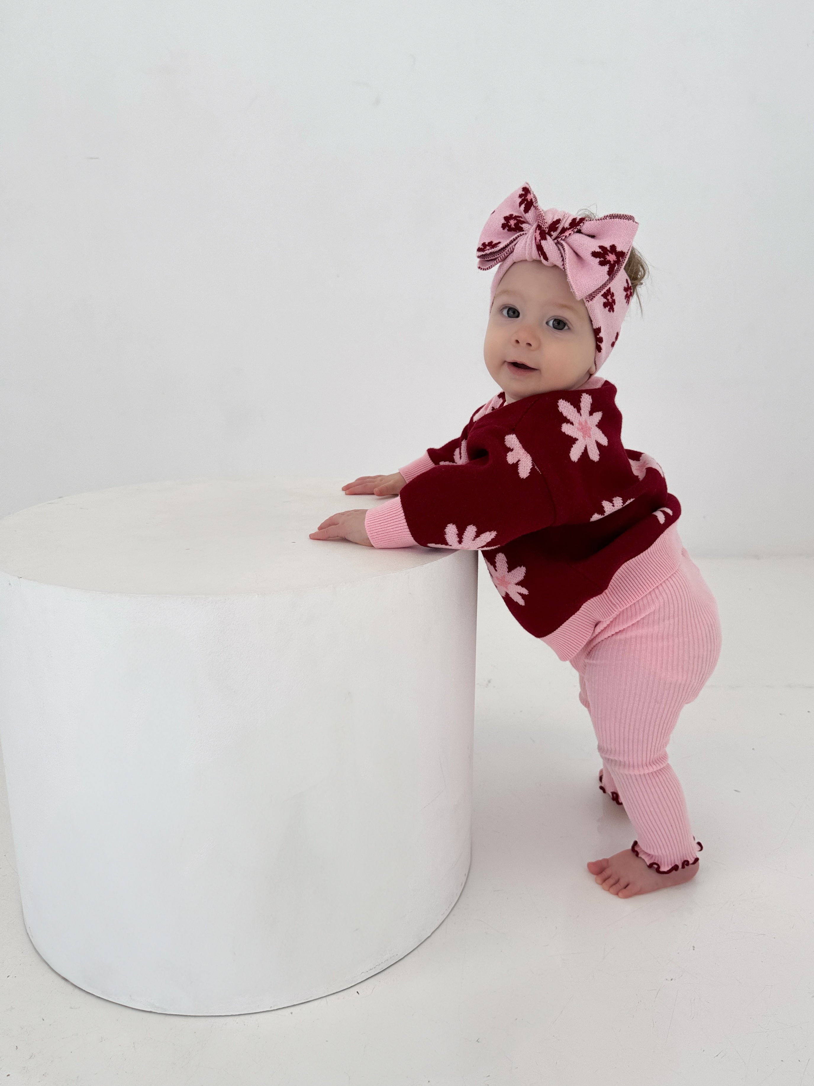 A baby in Ziggy Lou - Leggings Lola by ZIGGY LOU, a pink and red flower-patterned knitted legging with an elastic waistband and matching headband, stands barefoot holding a large white cylinder against a plain background.