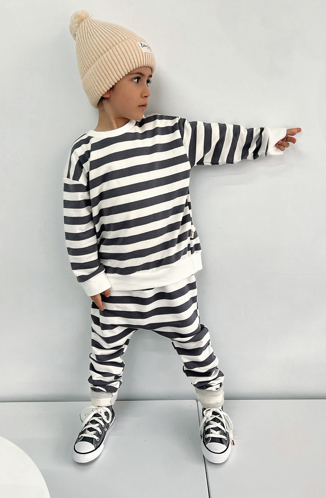 A young toddler wearing the Camden navy striped pullover and matching pants with the Bobby G cream beanie. He has his left arm stretched out and looking in the same direction. Hes wearing converse shoes.