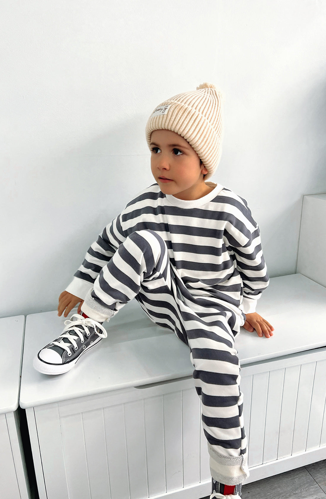 A young boy wearing the Camden navy striped pullover and matching sunny pants with the Bobby G creme beanie. He has one leg up on the seat.