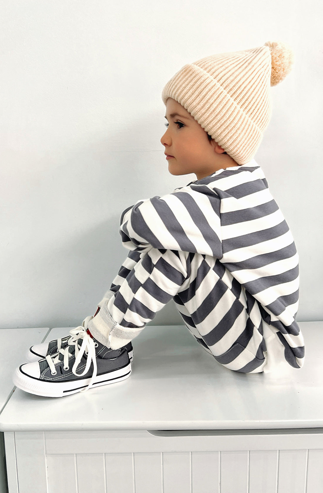 A young toddler wearing the Camden navy striped pullover and matching pants with the Bobby G cream beanie. He has his feet up on the bench and knees to his chin on a side on angle.