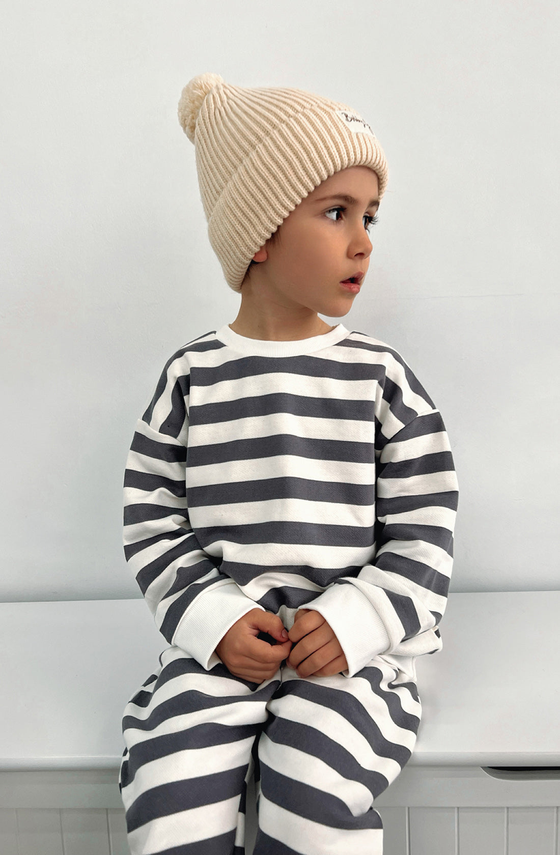 A young toddler wearing the Camden navy striped pullover and matching pants with the Bobby G cream beanie sitting on a white seat with their head turned.