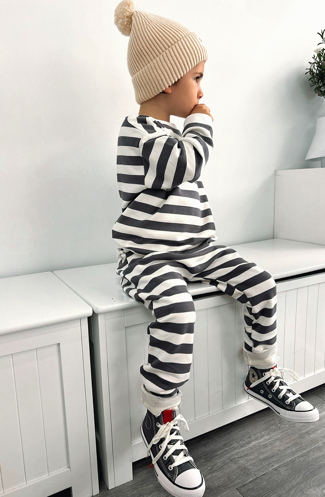 A young boy wearing the Camden navy striped pullover and matching pants with the Bobby G cream beanie. He's sitting on a seat facing his left.