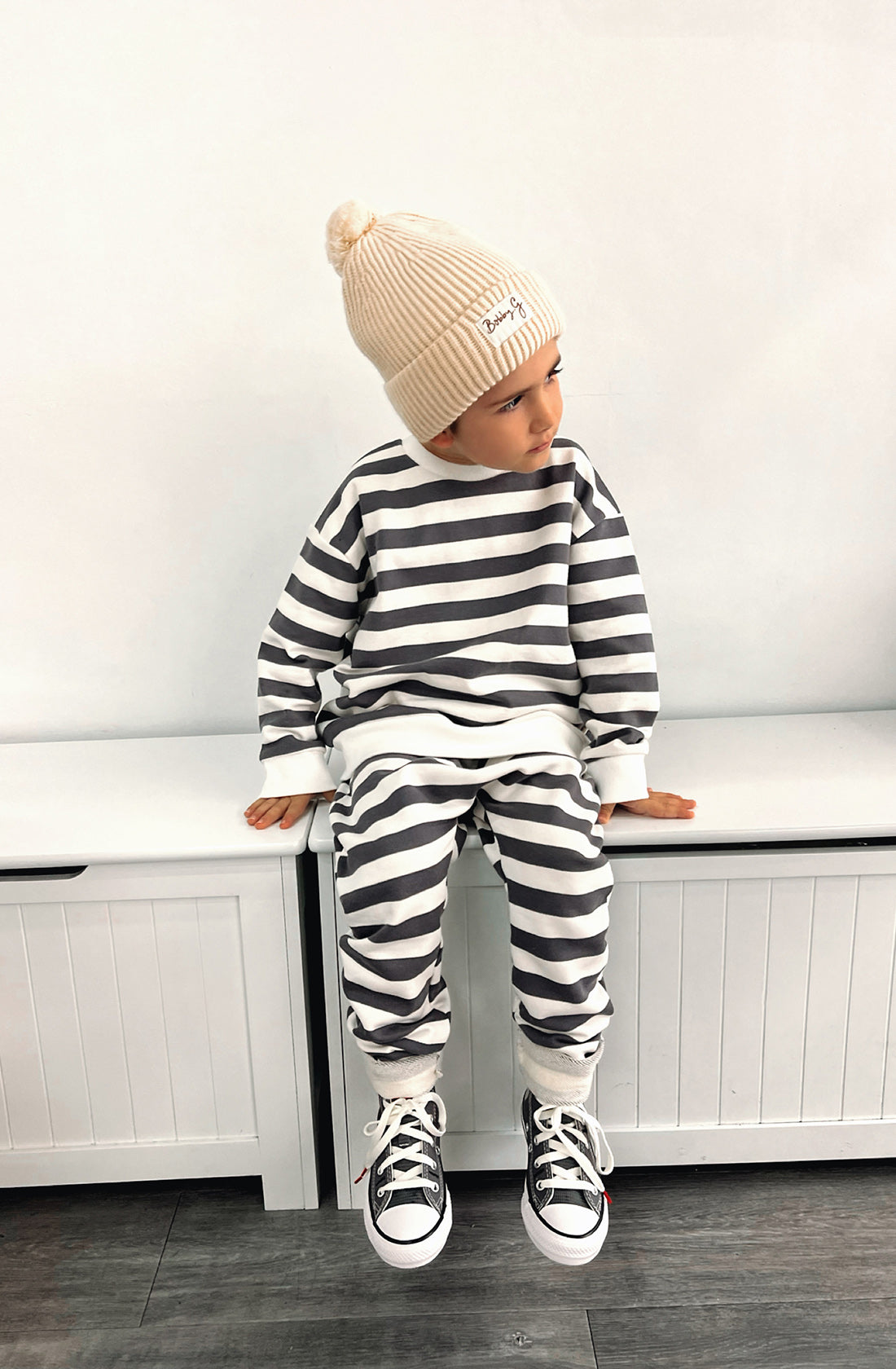 A young toddler wearing the Camden navy striped pullover and matching pants with the Bobby G cream beanie. He's sitting on a seat looking to his left.