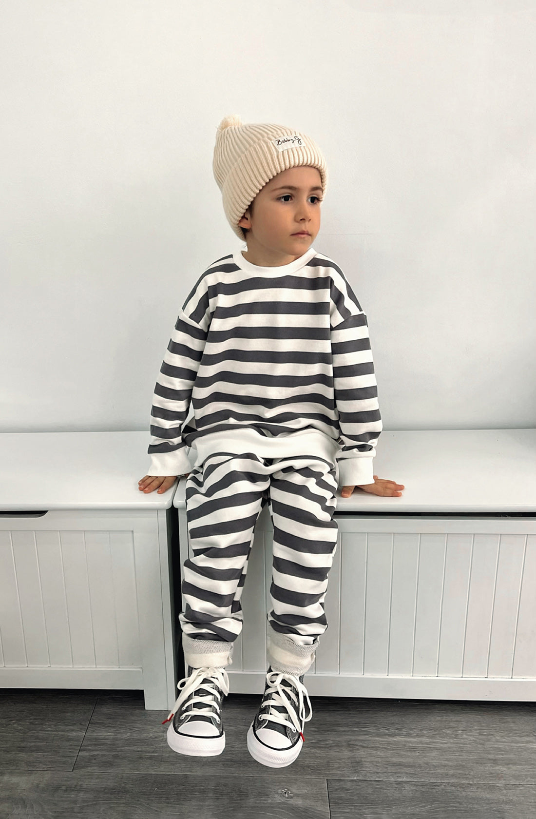 A young toddler wearing the Camden navy striped pullover and matching pants with the Bobby G cream beanie. He's sitting on a seat looking to his left.