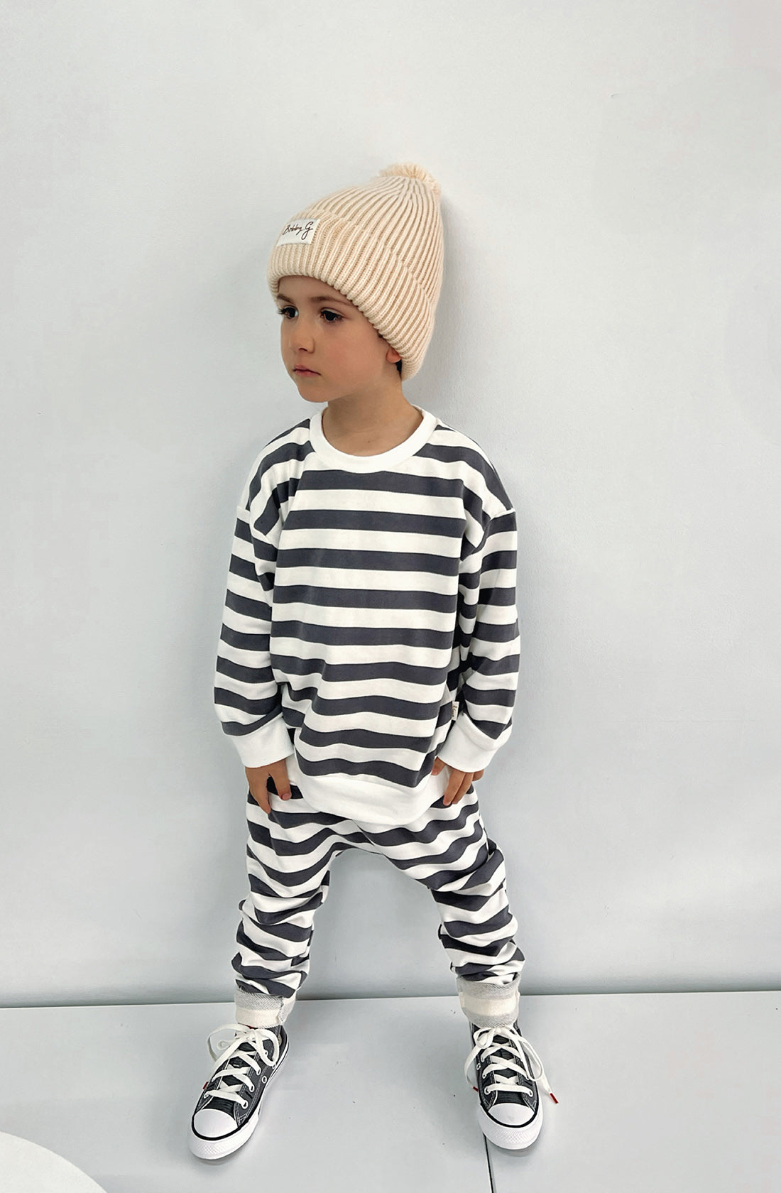 A young toddler wearing the Camden navy striped pullover and matching pants with the Bobby G cream beanie. He's looking to his right.