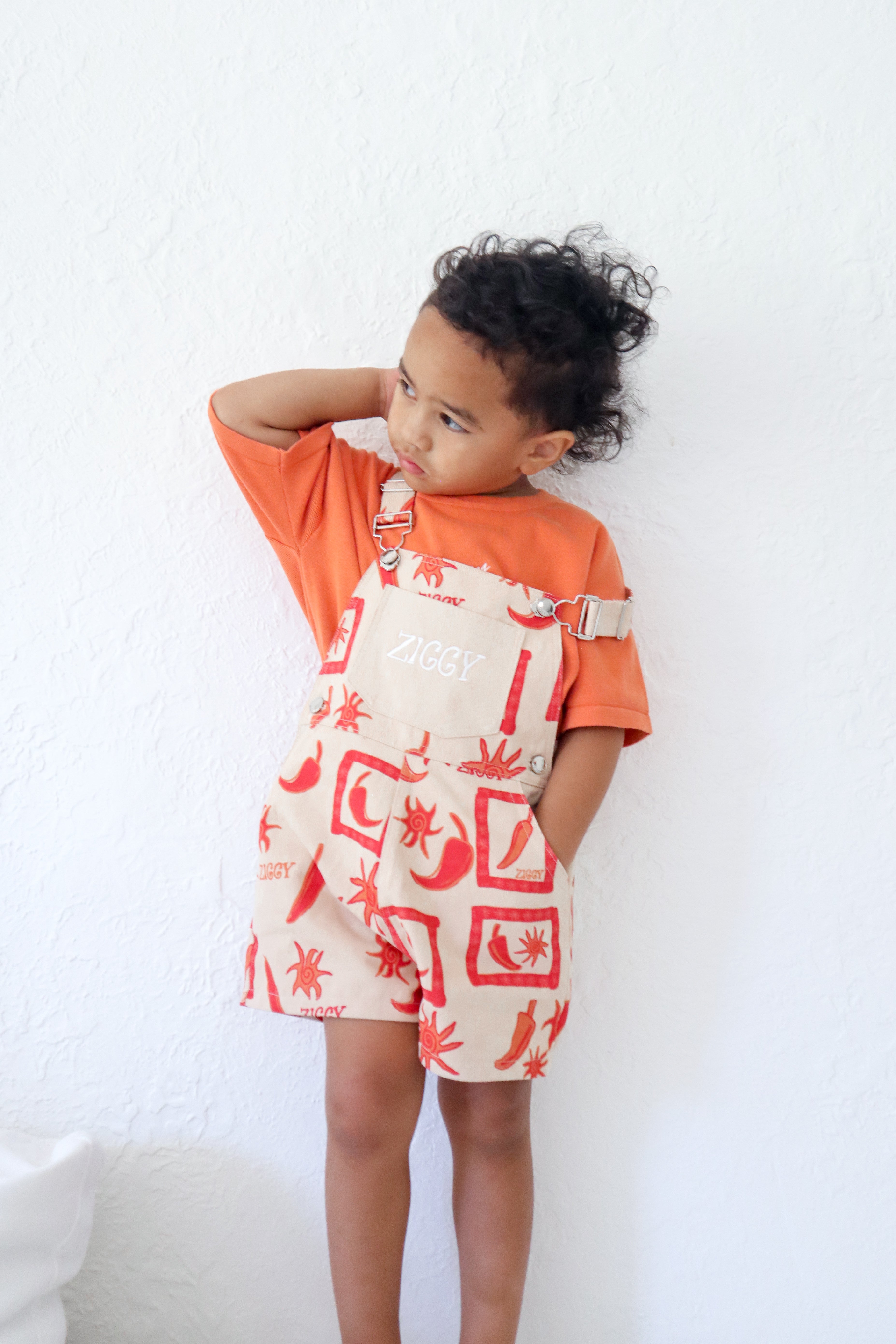 A young child with curly hair stands against a white wall, wearing Ziggy Lou ~ Short Overalls Chilli by ZIGGY LOU. The orange shirt and cream-colored overalls feature a vibrant chili design with adjustable straps for comfort. The child looks to the side curiously, hand behind head.