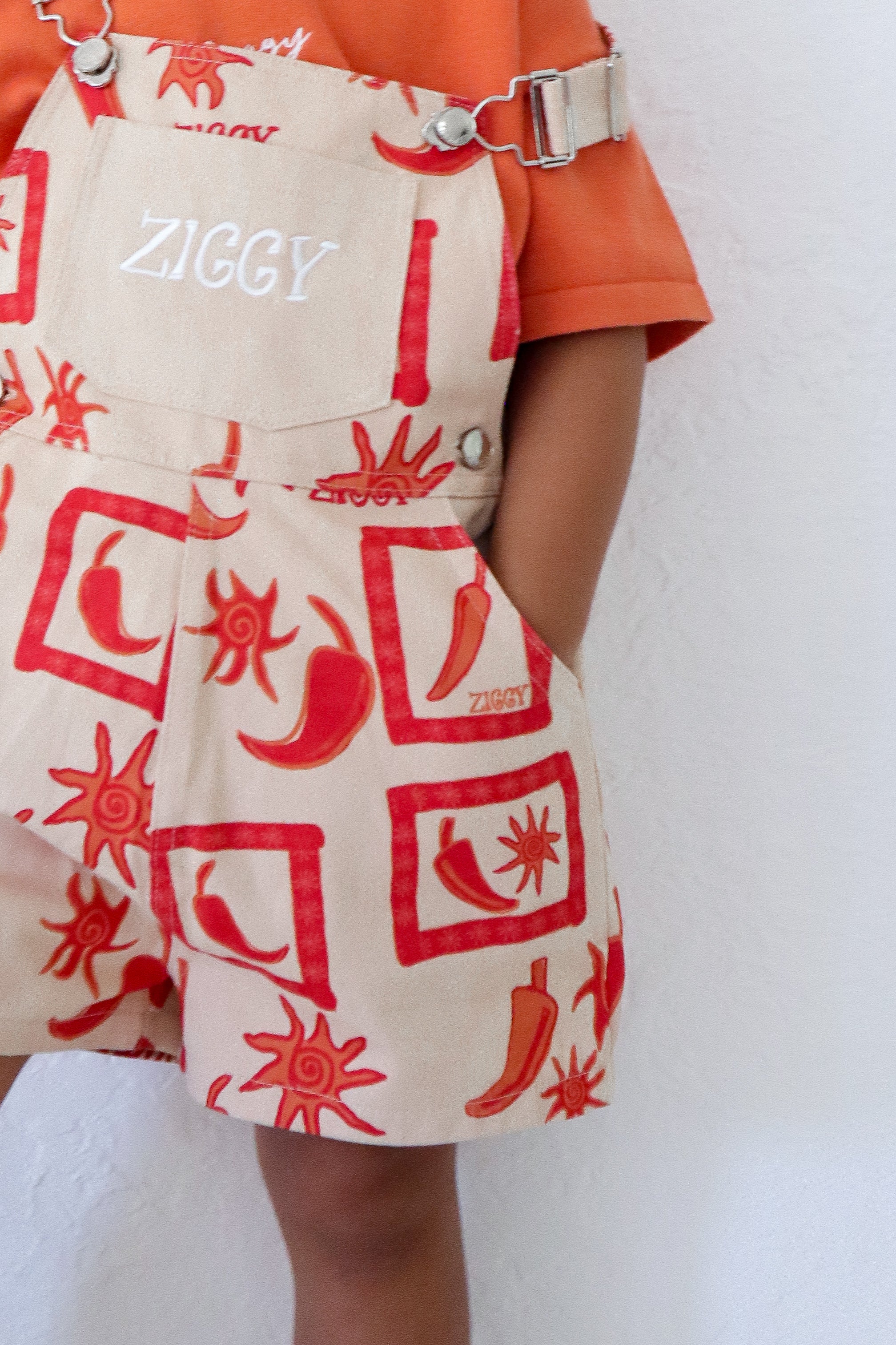 A child in Ziggy Lou short overalls with a chili design and sun patterns, "Ziggy" on the front, adjustable straps, and an orange shirt, casually stands with one hand in their pocket against a white backdrop.