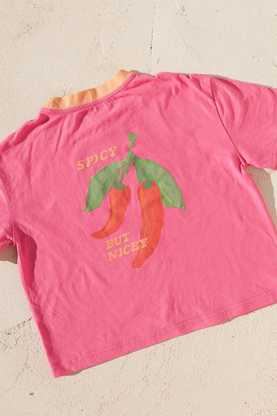 The SMALL SWIM CLUB Spicy But Nicey T-Shirt in Pink/Red features a Tex Mex-inspired design with an illustration of two chili peppers and the phrase "SPICY BUT NICEY" on the back. The green stems and leaves stand out vividly against the light pink backdrop, making it suitable for anyone. When laid flat on a light surface, it exudes vibrant western vibes flawlessly.