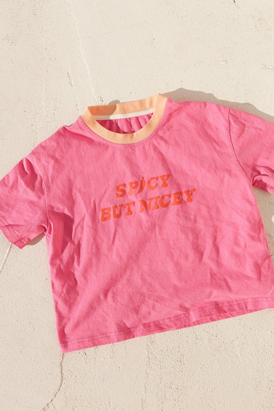 The Spicy But Nicey T-Shirt Pink/Red by SMALL SWIM CLUB, featuring bold orange letters and a Tex Mex flair, is displayed on a light-colored surface.