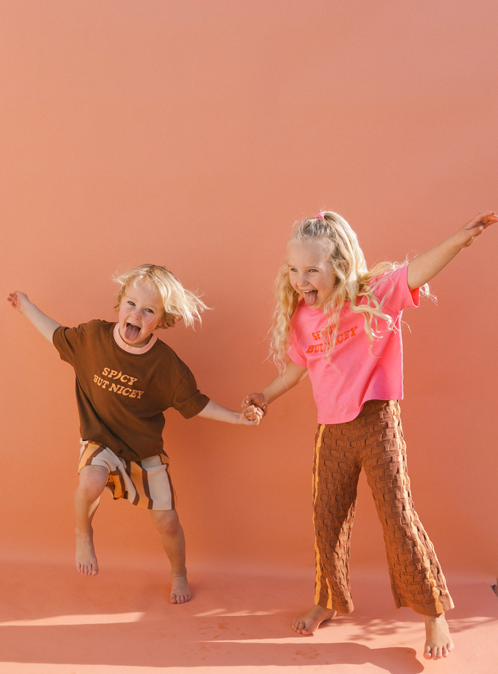 Two children jump joyfully, holding hands against an orange backdrop, exuding western vibes. The child on the left wears a brown t-shirt and striped shorts, while the one on the right sports a Spicy But Nicey T-Shirt in Pink/Red by SMALL SWIM CLUB paired with textured brown pants. Both smile energetically, embodying unisex charm.