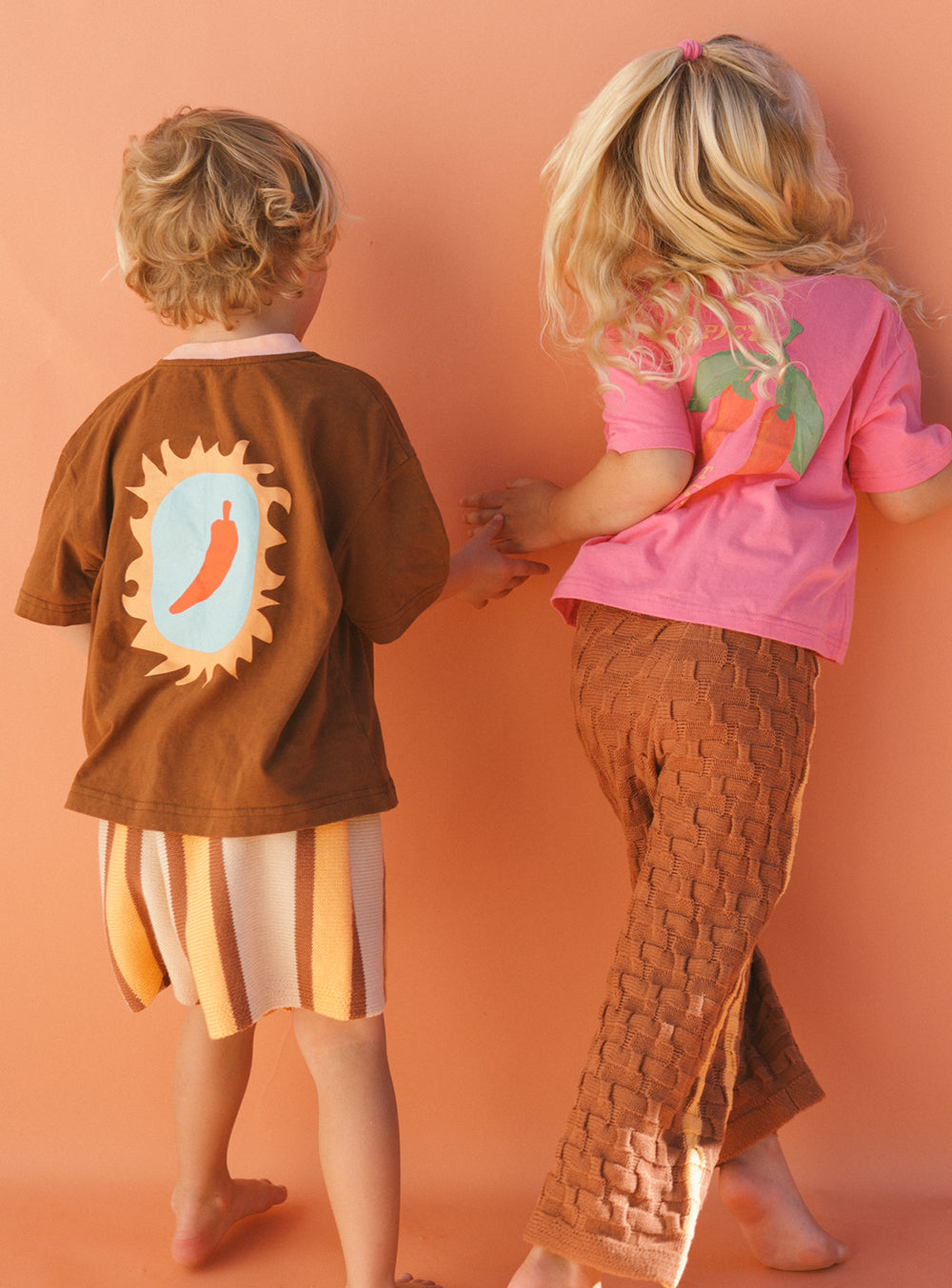 Two children are holding hands and facing away, showcasing a Tex-Mex style. One is dressed in the "Spicy But Nicey" T-shirt from SMALL SWIM CLUB, featuring a brown chili pepper design paired with striped shorts. The other wears a pink shirt with a carrot motif and brown woven pants against a solid peach background, enhancing their unisex appeal.