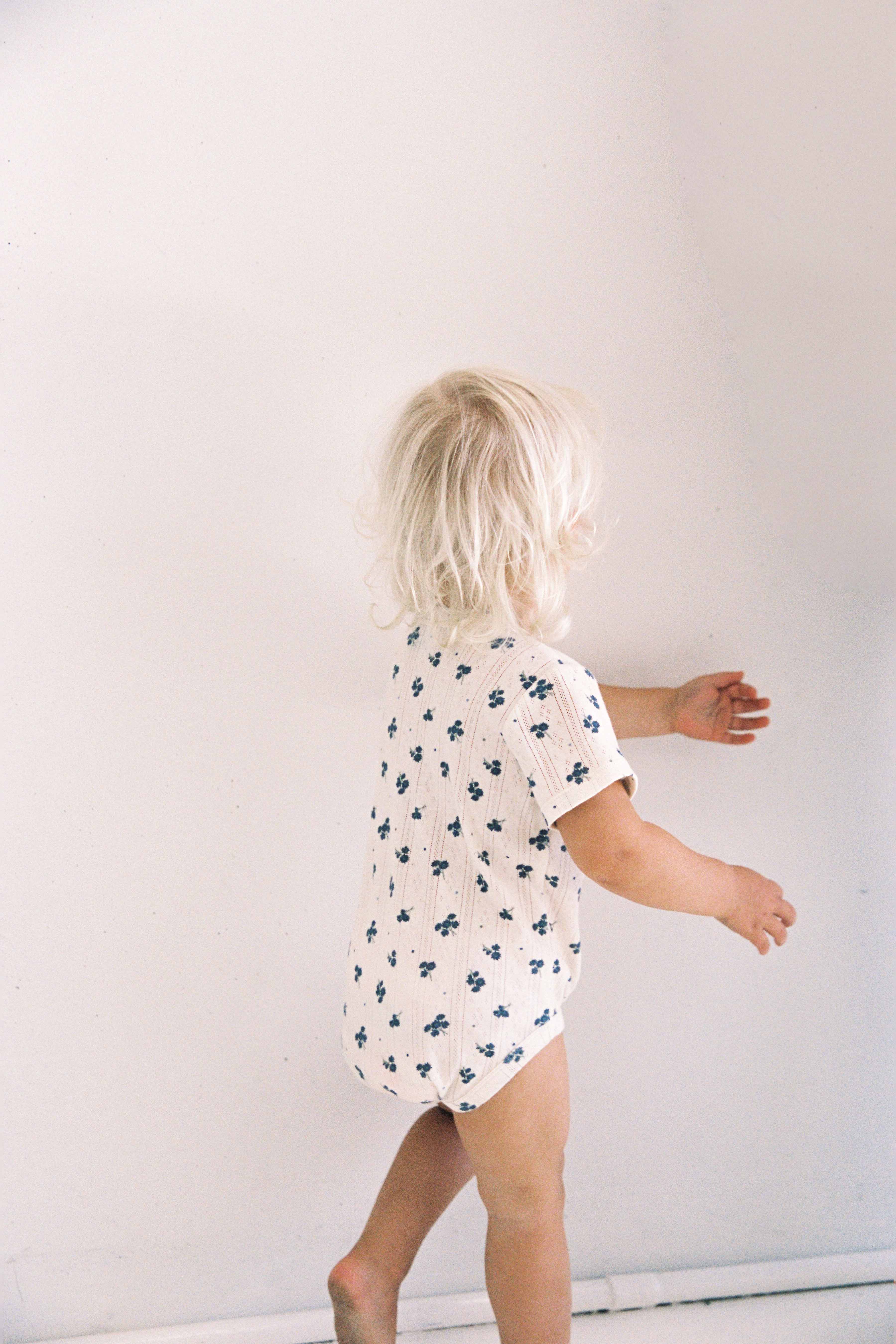 A blond toddler with a head full of wispy hair, wearing an ethically made Peony Pointelle Onesie by JUNI JUNIOR, stands against a light-colored wall and reaches out with their hands.