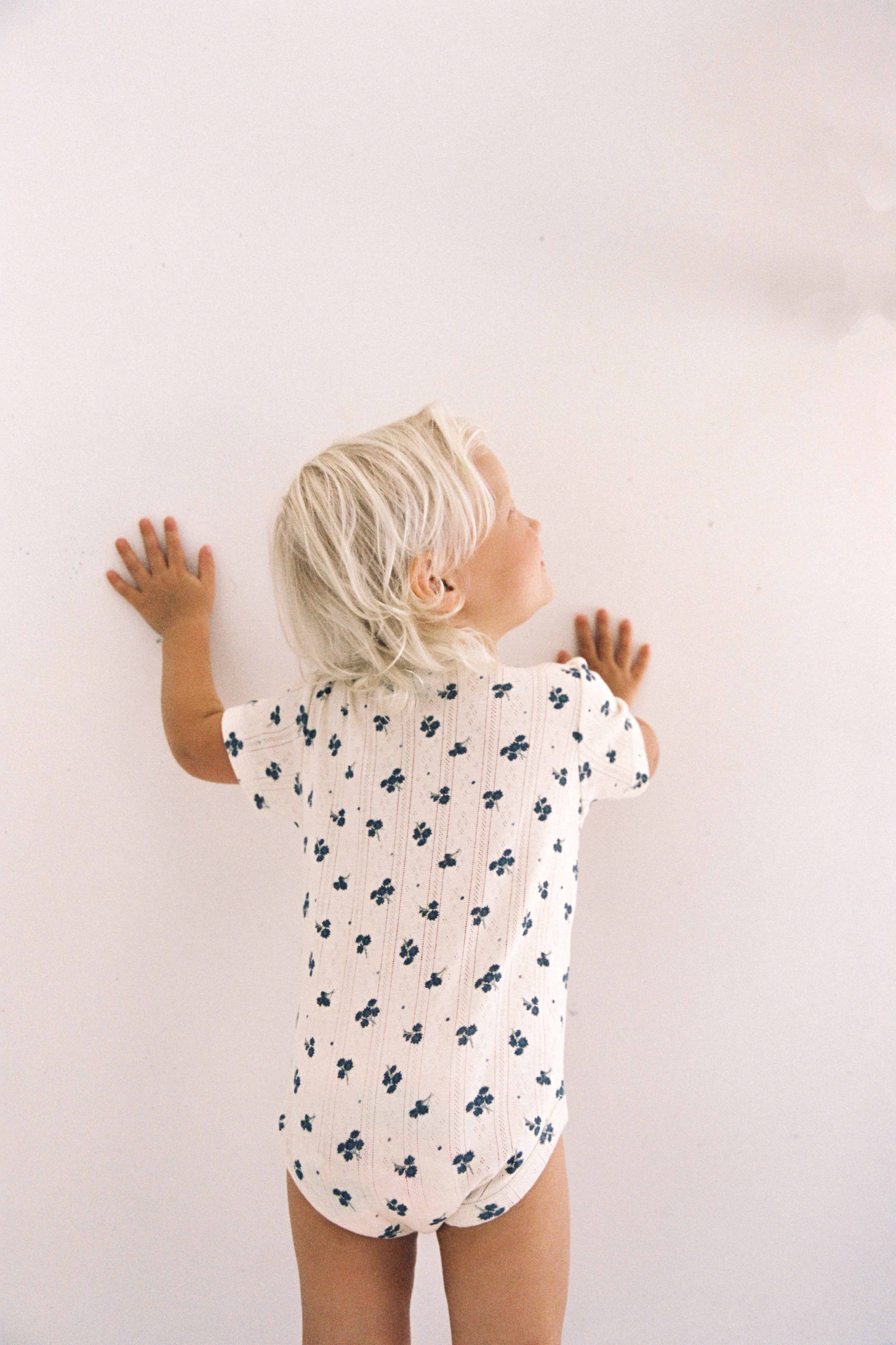 A young child with light blonde hair, seen from behind, stands against a plain white wall with their hands pressed against it. The child is wearing a JUNI JUNIOR Peony Pointelle Onesie made from ethically produced cotton, featuring small blue flower patterns on the short-sleeved white fabric.