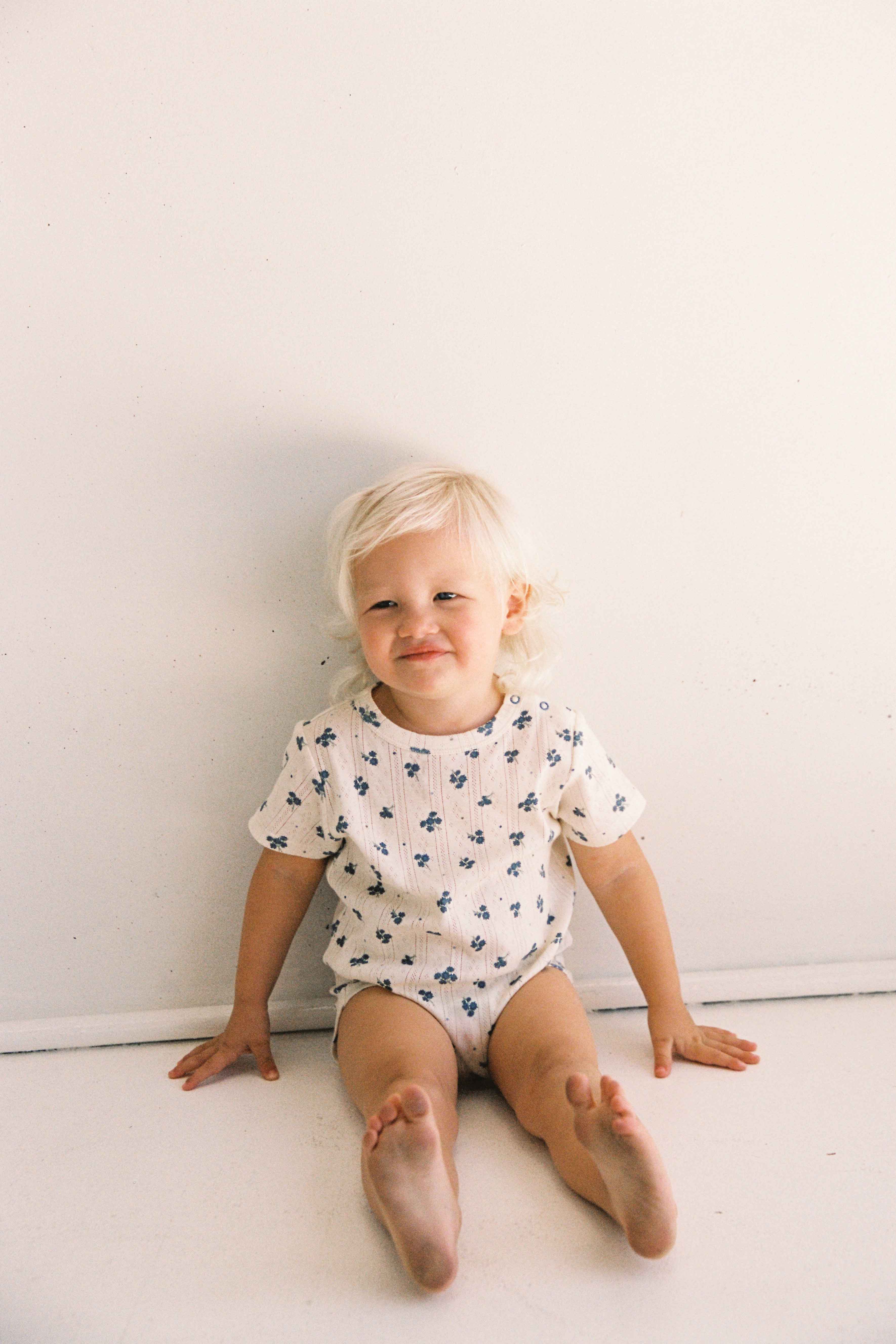 A young child with light blonde hair sits against a plain white wall, wearing the Peony Pointelle Onesie from JUNI JUNIOR, adorned with small blue floral patterns. The ethically made cotton fabric enhances the child's contented smile and peaceful demeanor.