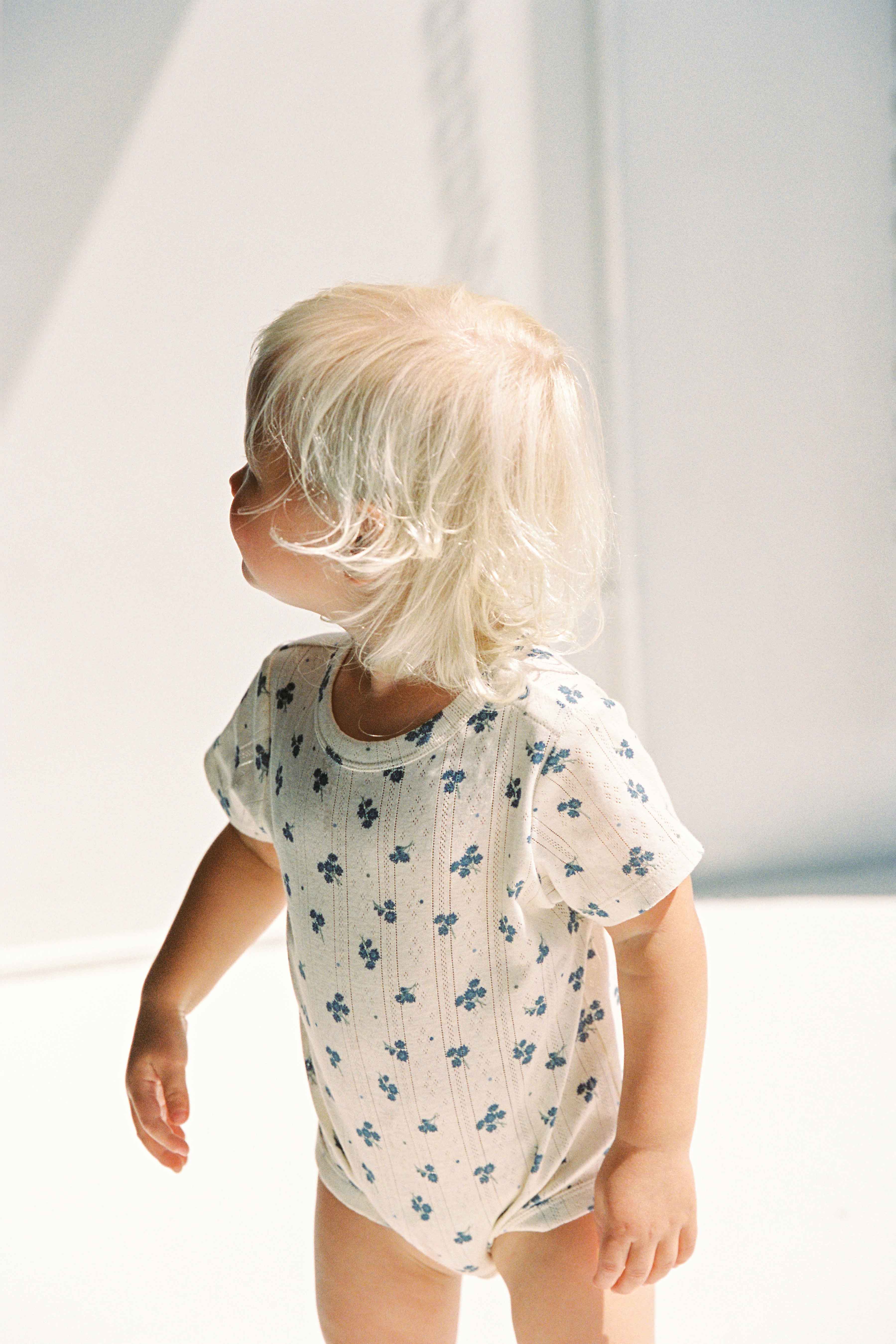 A small child with light blonde hair stands and looks to the side, wearing a Peony Pointelle Onesie from JUNI JUNIOR. The onesie, made from ethically sourced cotton pointelle jersey, features delicate blue floral patterns. The background is bright, with soft shadows suggesting natural light.