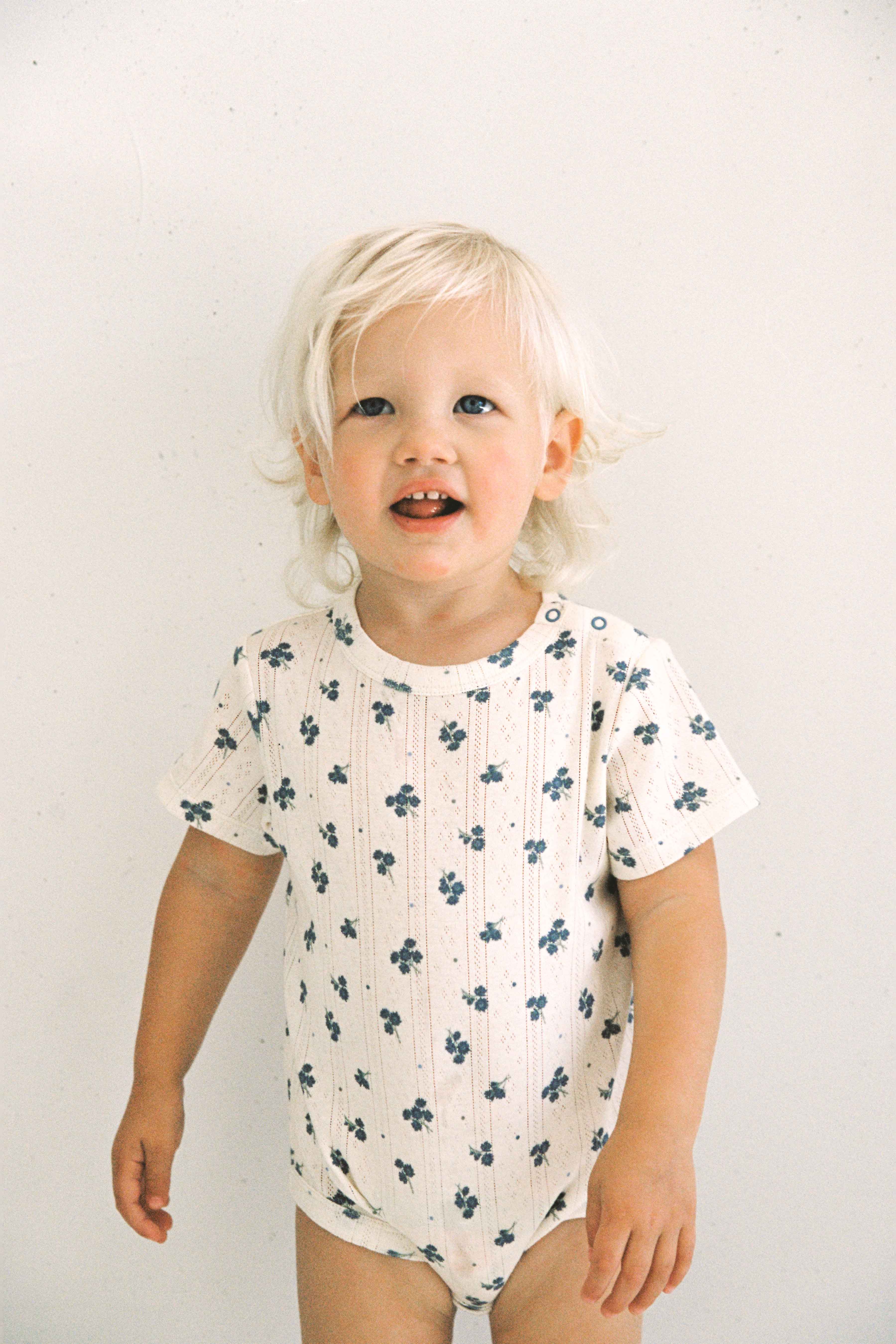 A toddler with light blonde hair is standing and smiling. They are wearing the Peony Pointelle Onesie by JUNI JUNIOR, a short-sleeved white jersey onesie featuring a small blue floral pattern made from soft cotton. The background is plain and light-colored.