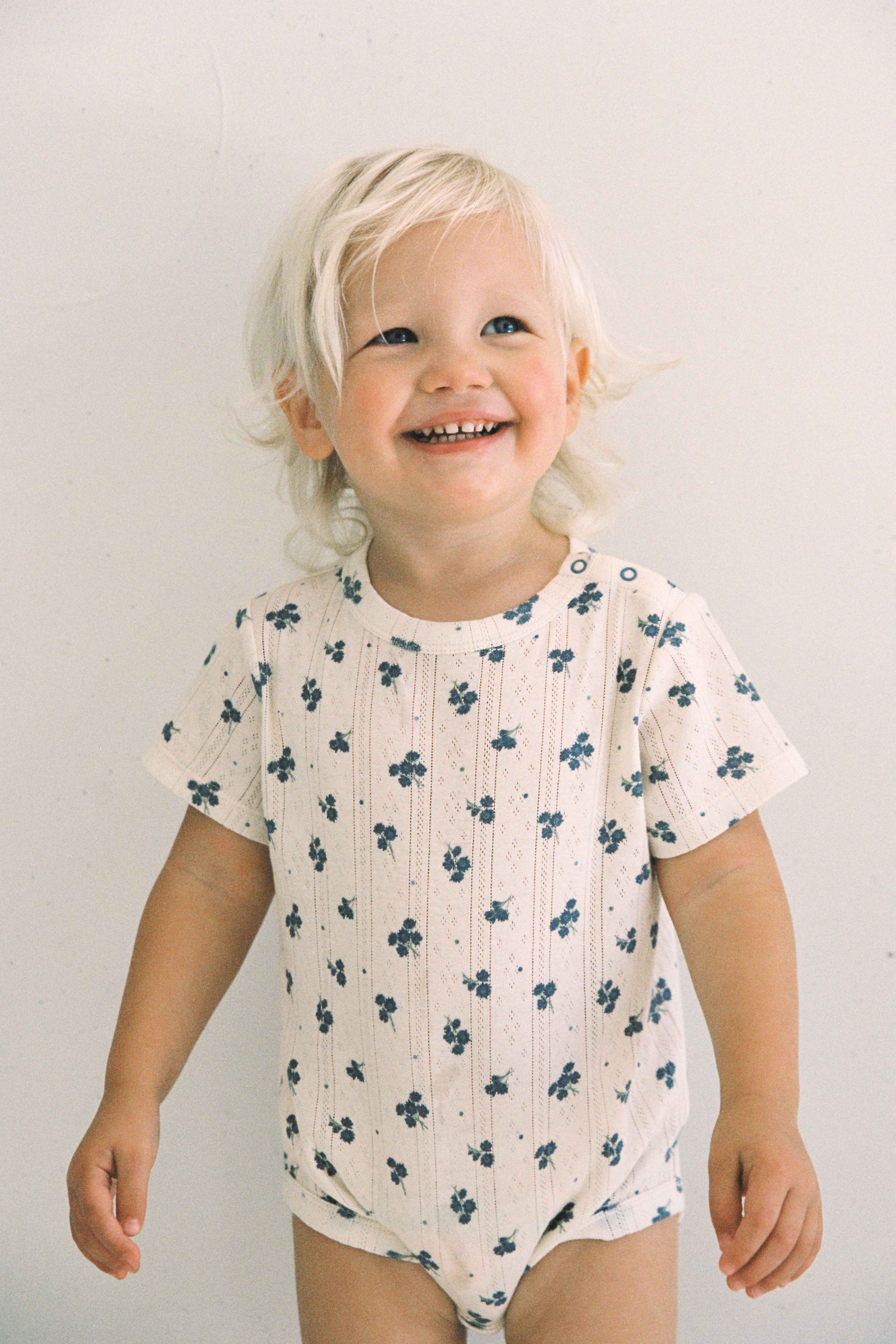 A joyful toddler with light blonde hair stands against a plain background, dressed in the Peony Pointelle Onesie from JUNI JUNIOR. The ethically made cotton onesie features small blue floral patterns. The child smiles brightly, and their hands rest relaxed by their sides.