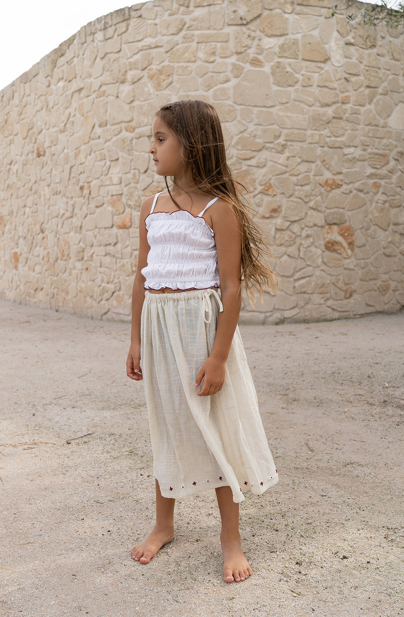 Girl wearing mid length natural skirt with white shirred crop top. Standing against a curved sandstone wall.