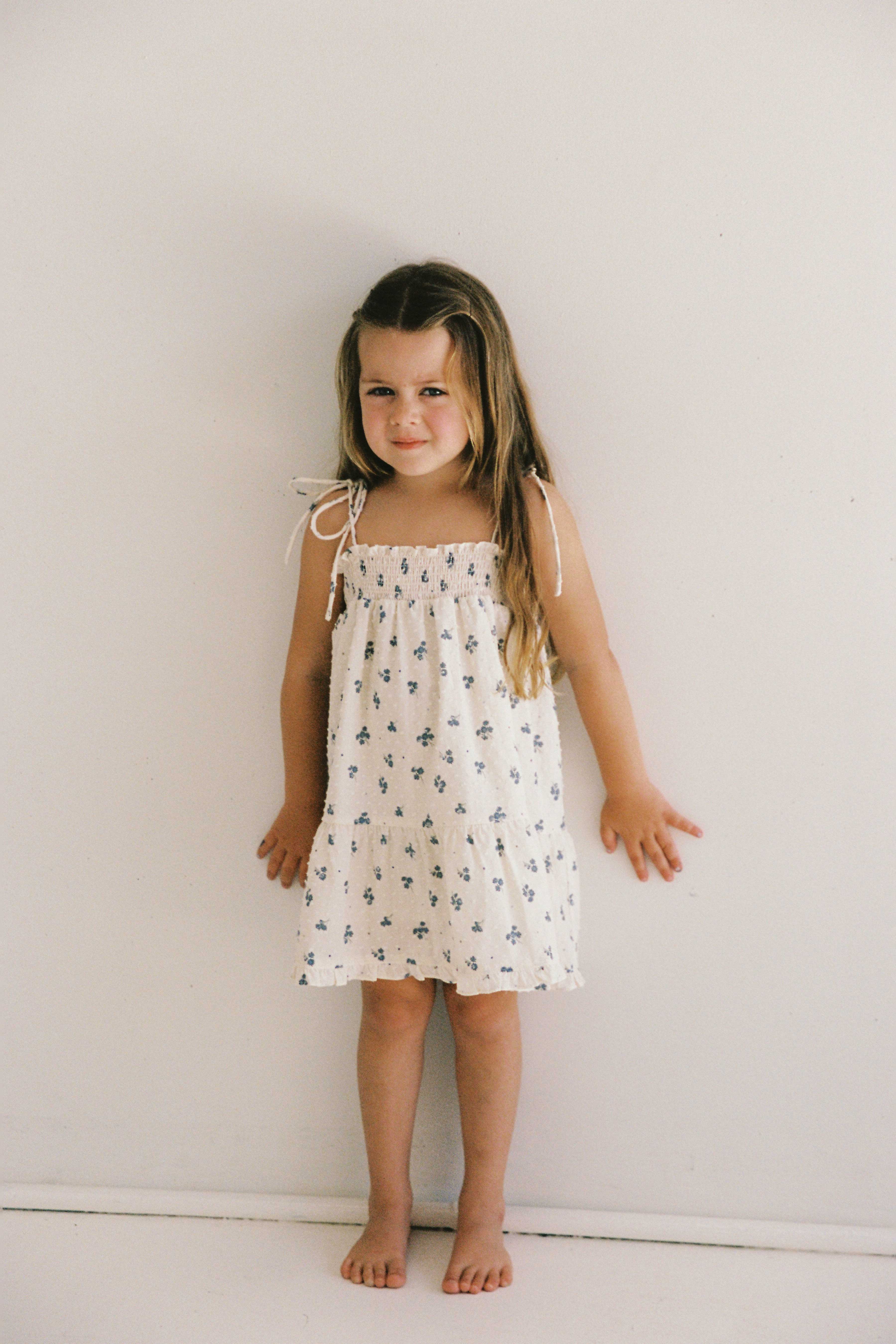 A young girl with long brown hair stands barefoot against a plain white wall. She is wearing the sleeveless Posy Dress Ditsy Floral from JUNI JUNIOR, featuring a blue floral pattern and adjustable straps. The textured cotton dobby fabric of the ethically made dress adds charm to her outfit. Her hands rest gently against the wall, and she has a slight smile on her face.