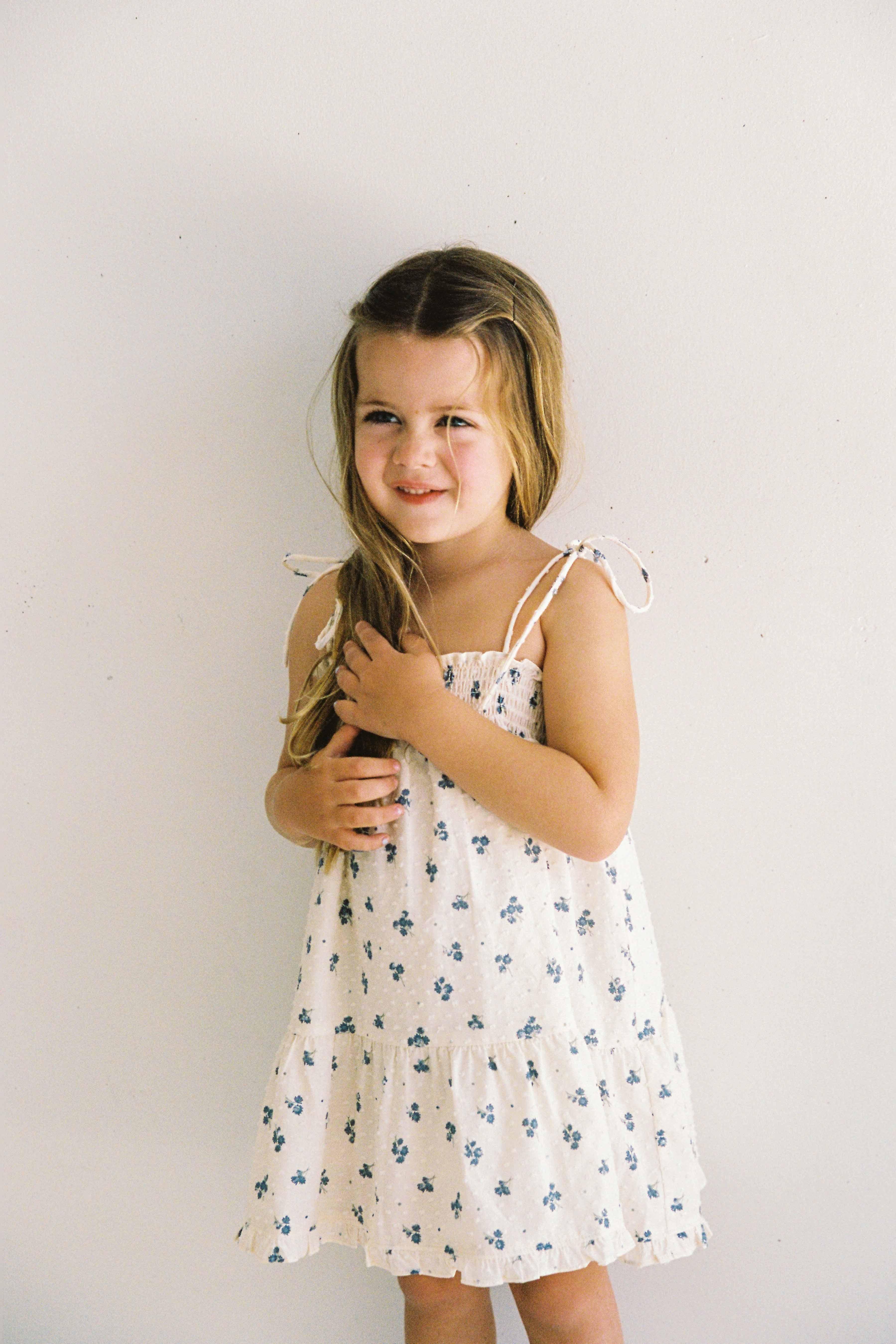 A young girl with long blond hair is standing against a plain white background, wearing the Posy Dress Ditsy Floral from JUNI JUNIOR. The dress features blue floral patterns and adjustable straps. She is smiling and holding part of her hair in one hand, effortlessly showcasing the textured cotton dobby fabric.