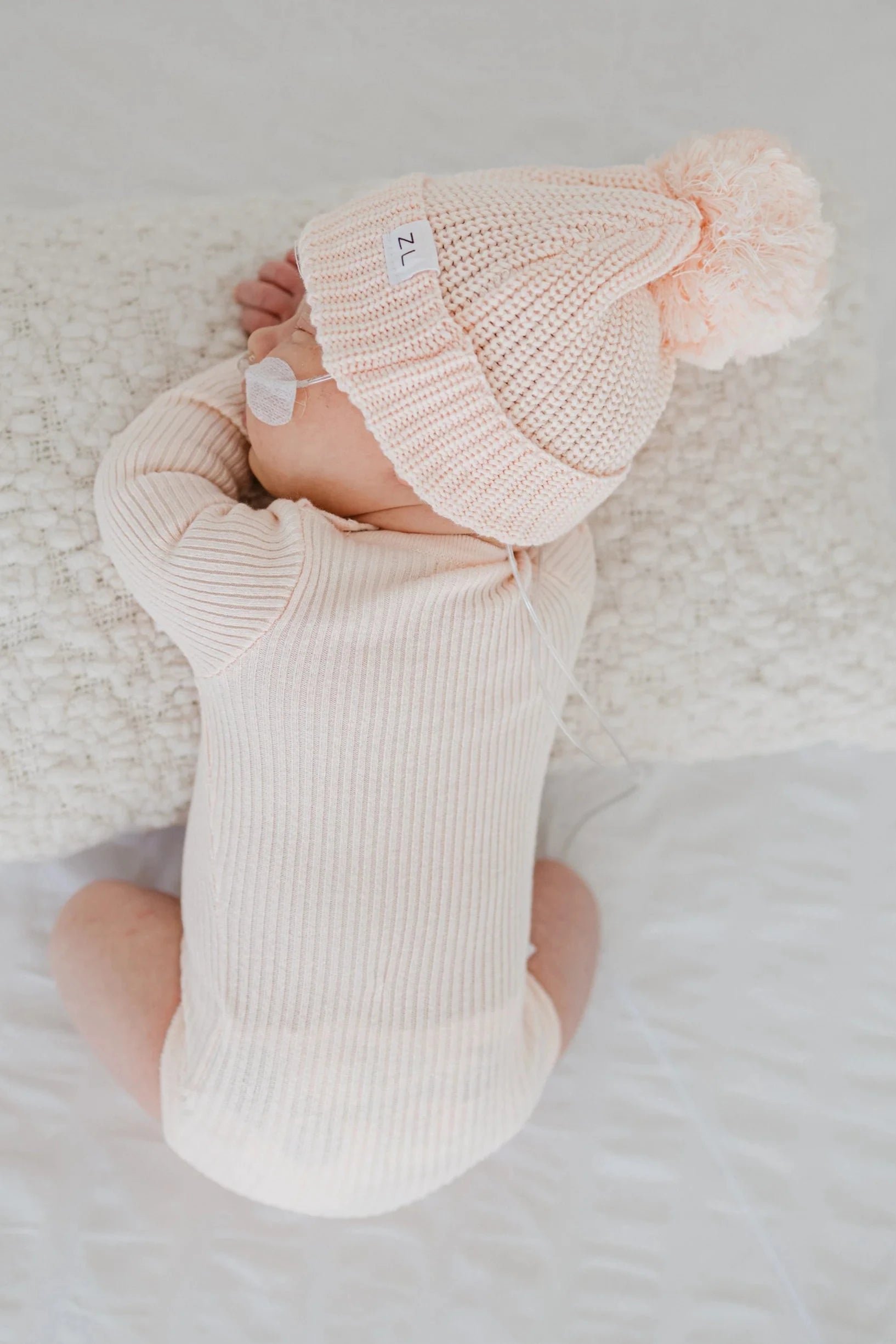 A baby wearing a ZIGGY LOU Beanie Primrose, which is a light pink chunky knit beanie with a large pom-pom, along with a matching 100% cotton knitted onesie, lies on their stomach on a white blanket and uses a pacifier.