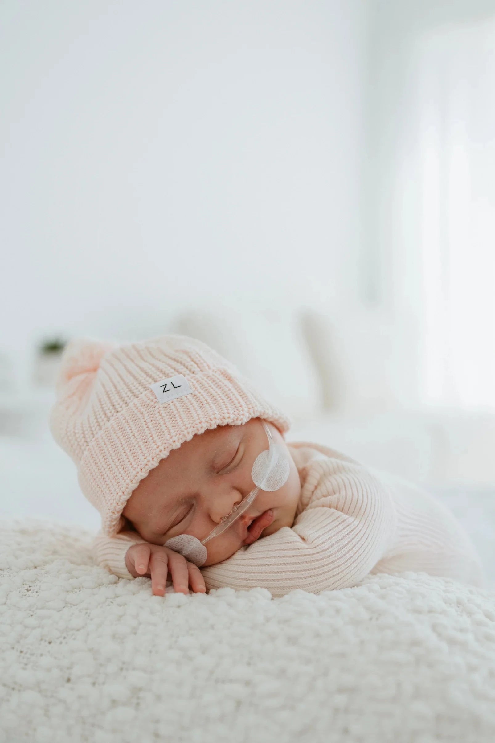 An infant peacefully sleeping on a soft white surface, wearing a light pink Beanie Primrose from ZIGGY LOU and a matching onesie. A small medical tube is attached to the baby’s face. The background is softly lit and out of focus.