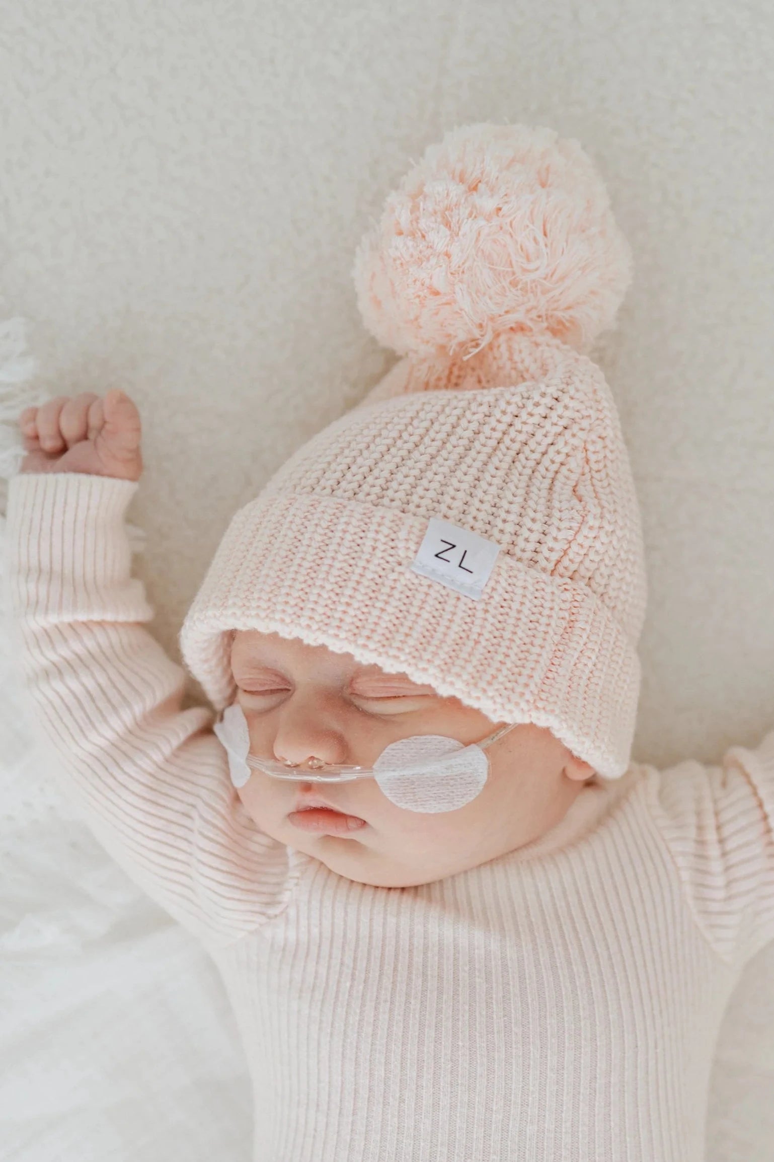 A sleeping baby wearing the ZIGGY LOU Beanie Primrose, a chunky knit beanie in pale pink with a large pom-pom and a white patch featuring the letters "ZL." The baby also has small medical adhesive patches and a thin nasal tube on its face, dressed in a cream-colored, ribbed long-sleeve outfit made of 100% cotton.