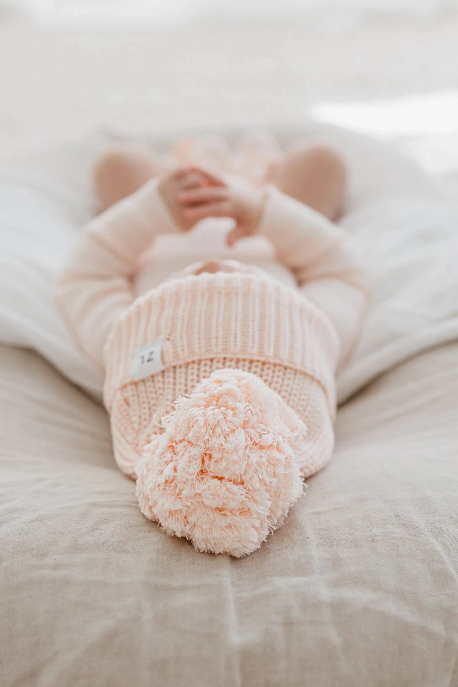 A baby wearing the ZIGGY LOU Beanie Primrose, a pink knitted outfit, and a chunky knit beanie is lying on a soft beige blanket. The baby is photographed from the feet up, with hands folded on the chest and the face mostly obscured by the Beanie Primrose, which features a large pom-pom and custom dyed aero fleck.